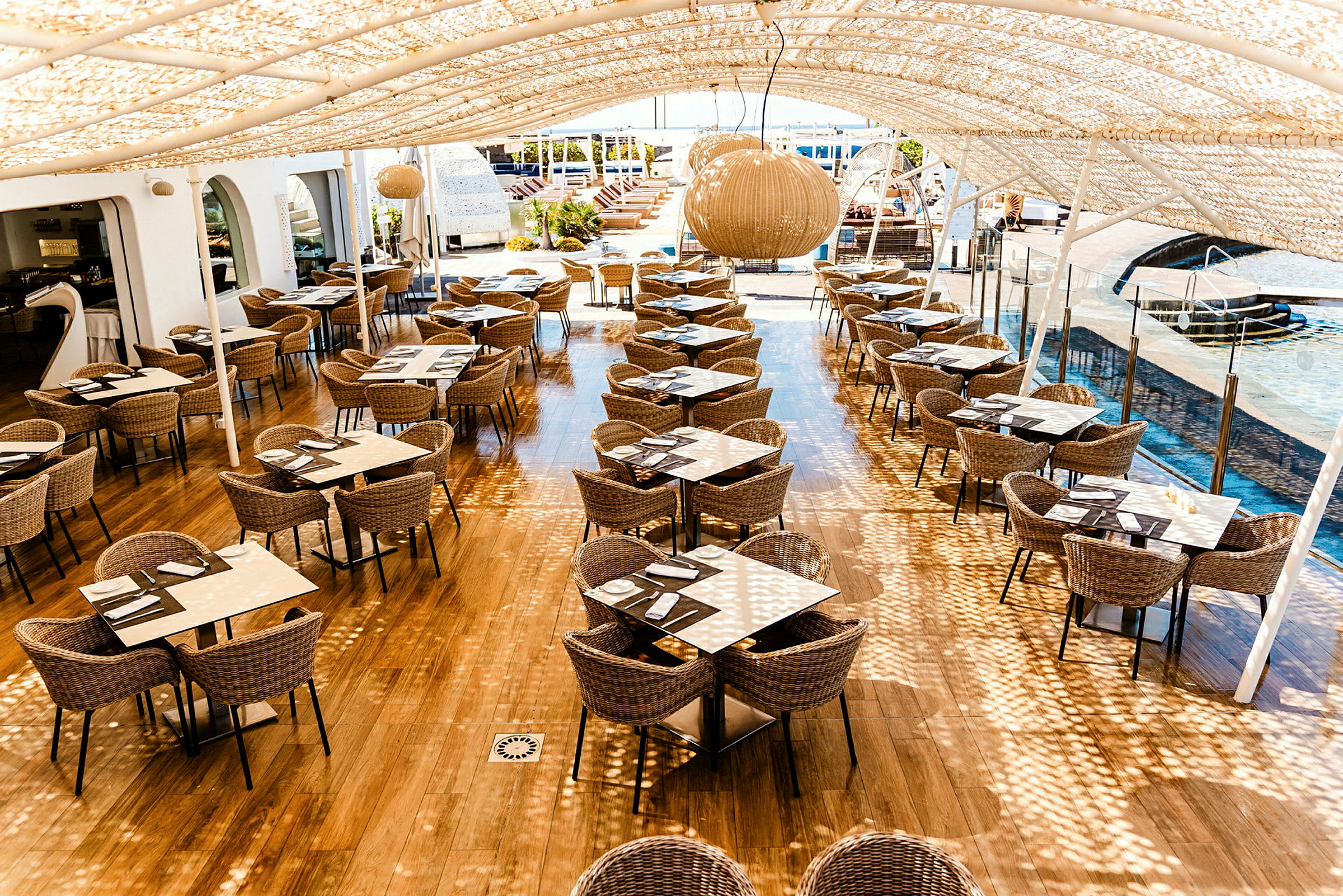 Utendørsrestaurant med skyggetak og sitteplasser ved bord av rotting og glass. (AI-generert tekst)