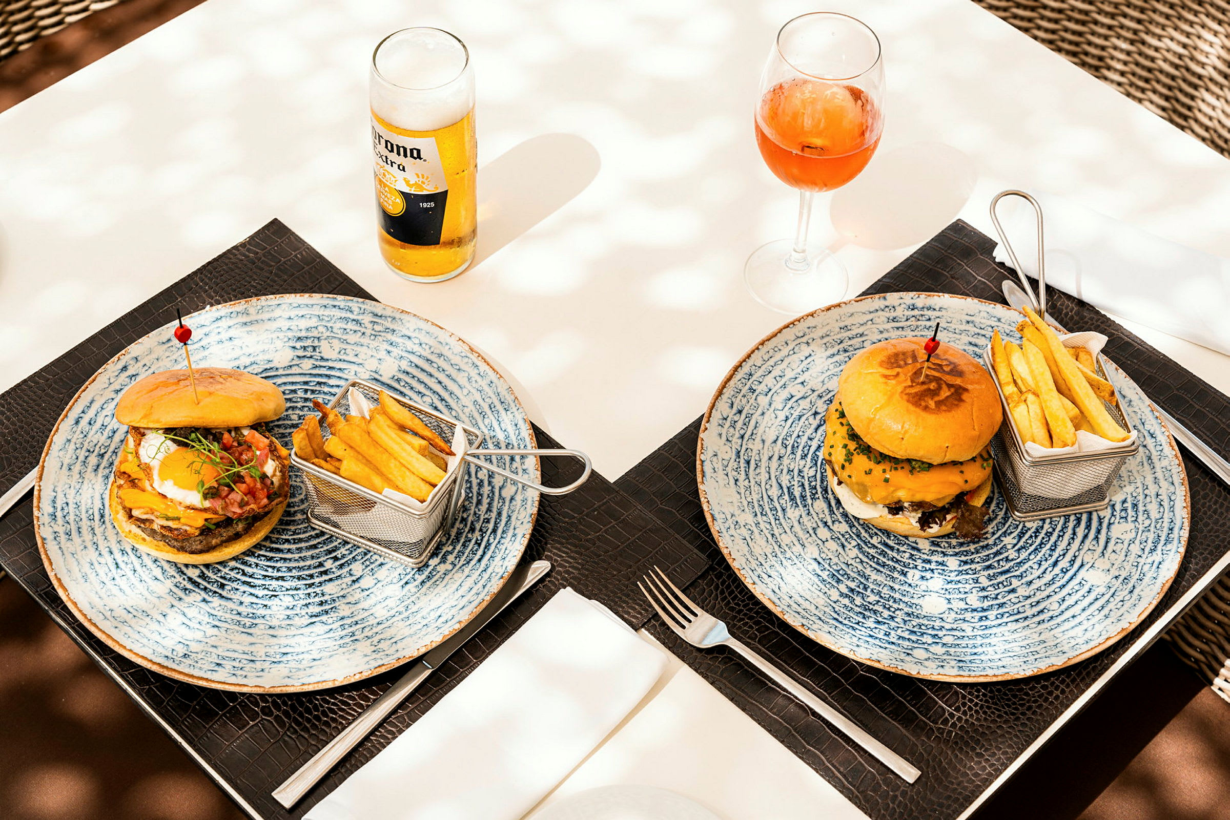 To tallerkener med hamburgere, pommes frites i små kurver, en øl og et glass med drikke på et dekket bord. (AI-generert tekst)