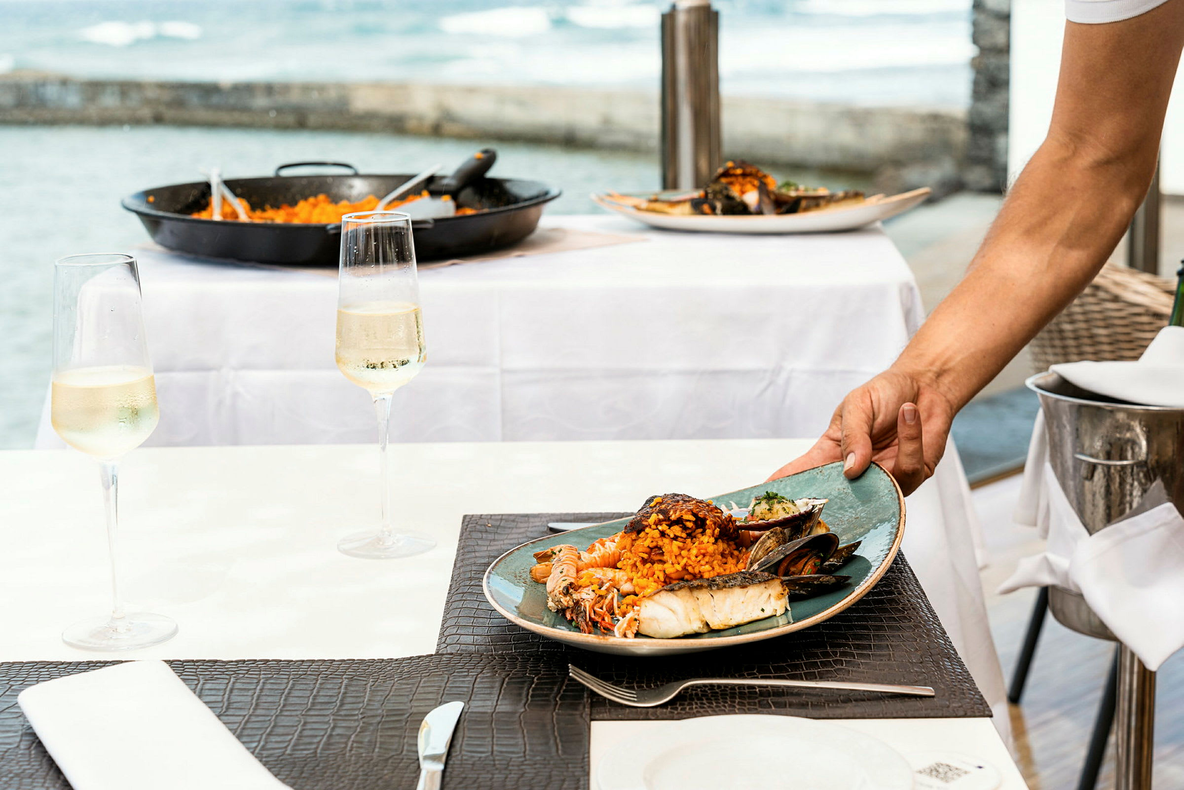 En tallerken med sjømat og ris serveres på et bord. En havutsikt er synlig bak bordet, og et glass med vin står på bordet. (AI-generert tekst)