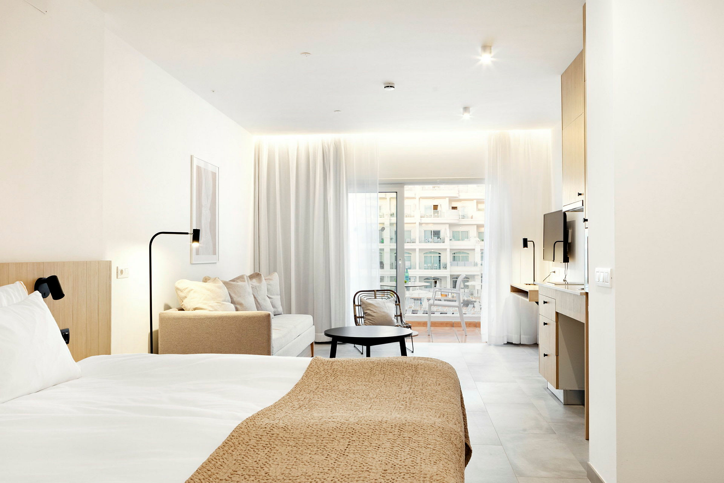 Moderne hotellrom med seng, sittegruppe og arbeidsområde, opplyst av naturlig lys fra balkongdører. (AI-generert tekst)