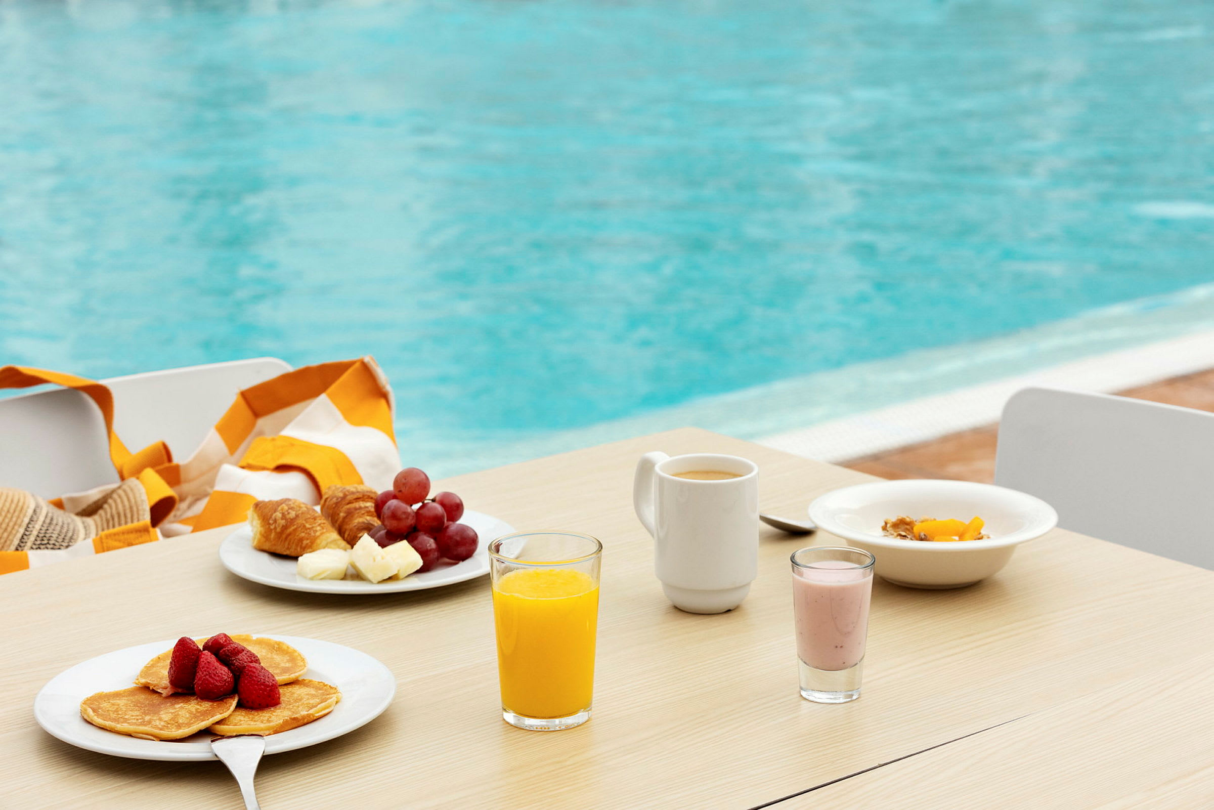 Frokost med frukt, pannekaker, juice og kaffe på et bord ved et basseng. (AI-generert tekst)