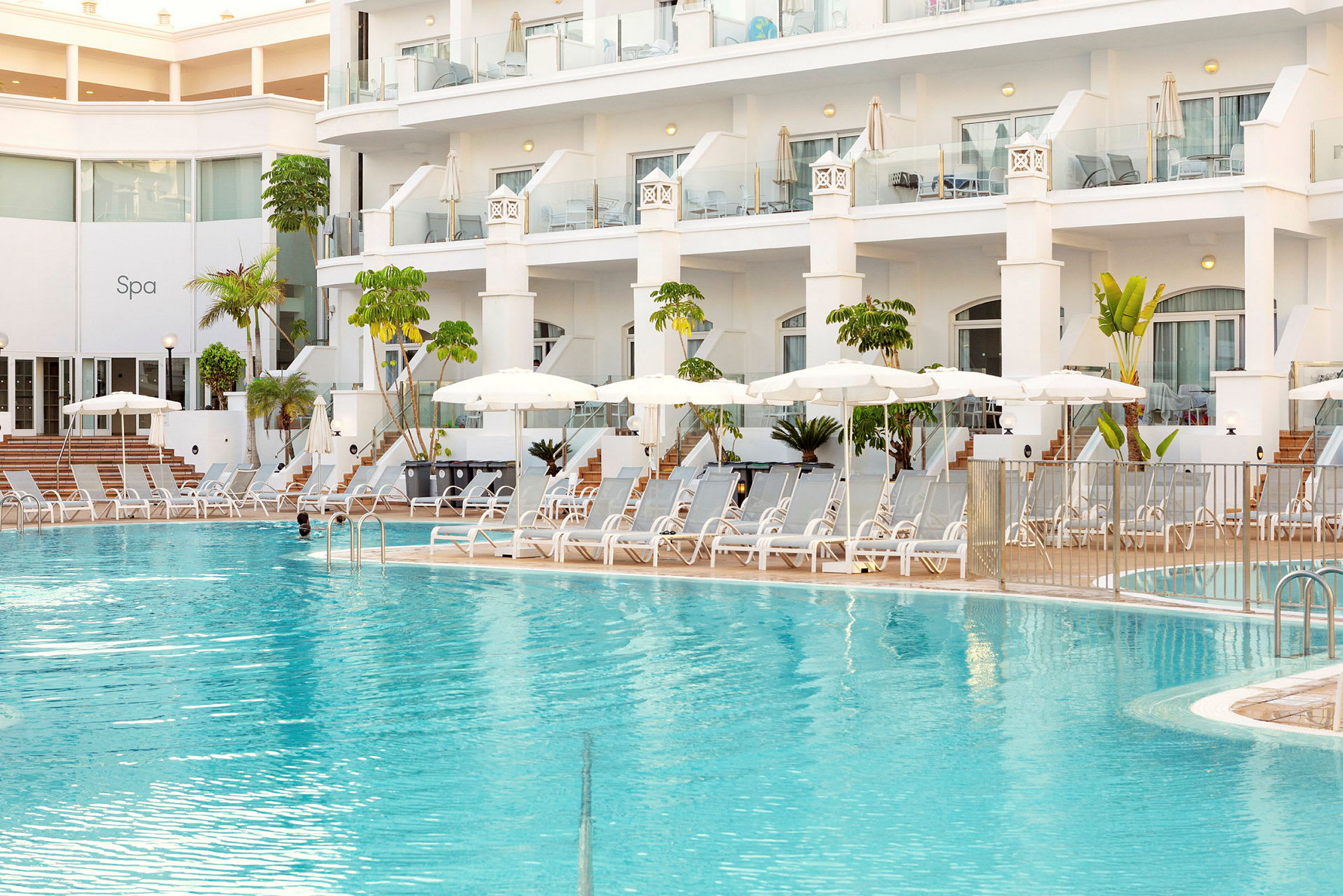 Hotellbasseng med solsenger og parasoller ved en moderne bygning. (AI-generert tekst)