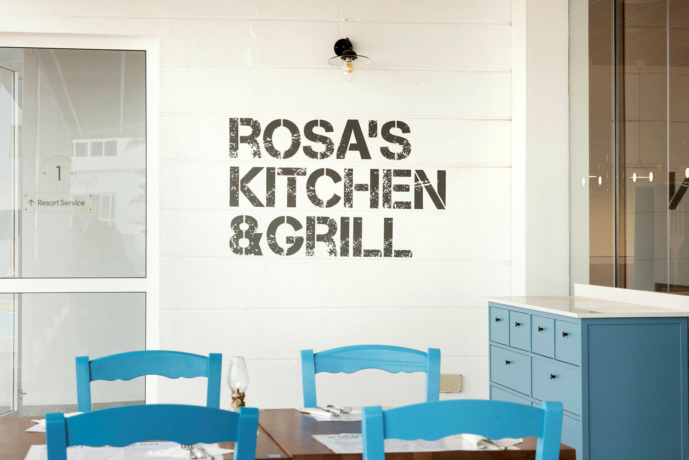 Skilt med teksten 'Rosa's Kitchen & Grill' på en vegg, bord og blå stoler i forgrunnen. (AI-generert tekst)