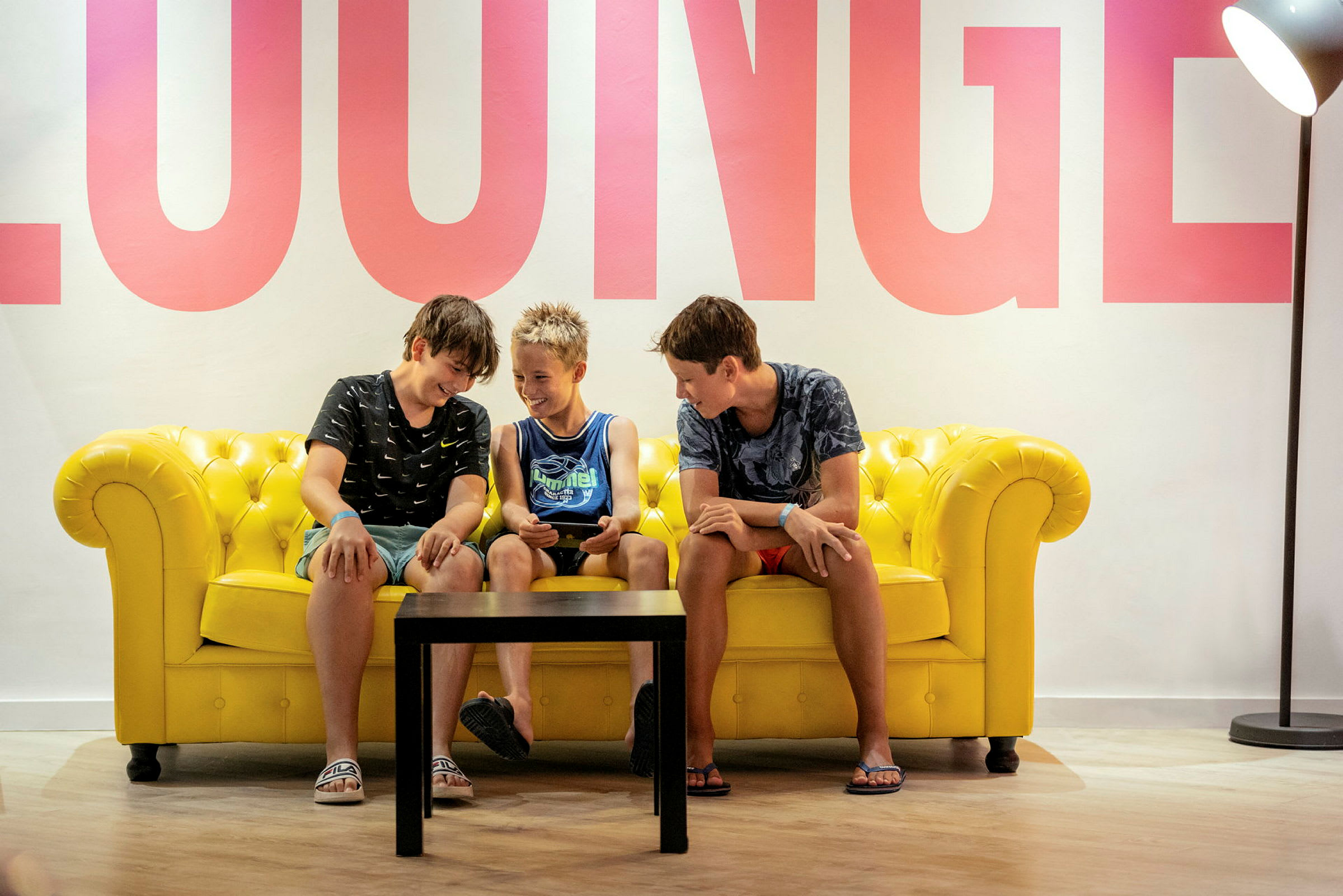 Personer sitter på en gul sofa med et bord foran, og ordet 'LOUNGE' er synlig på veggen. (AI-generert tekst)