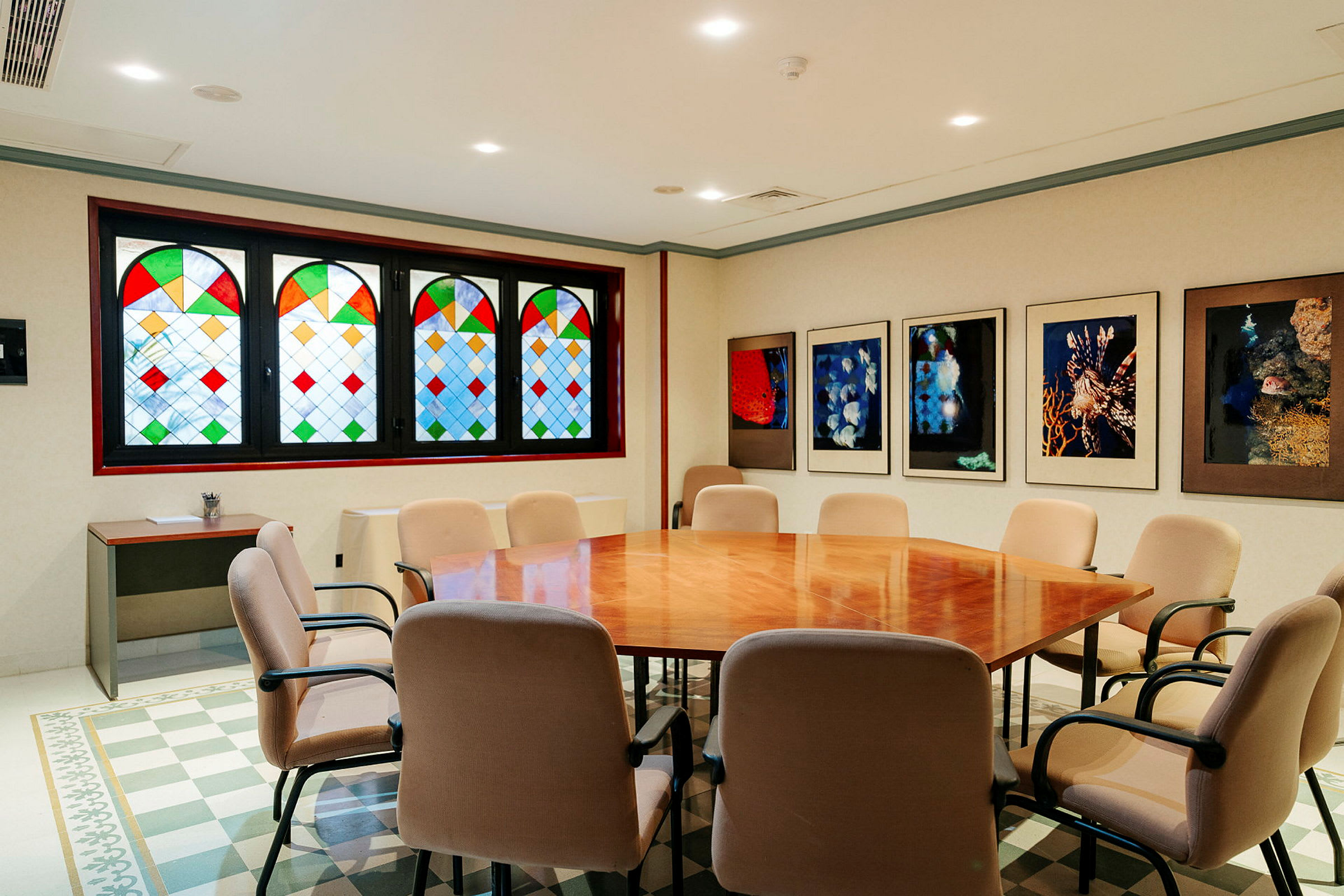 Et konferanserom med et stort trebord omgitt av stoler, vinduer med farget glass og vegger dekorert med kunstverk. (AI-generert tekst)