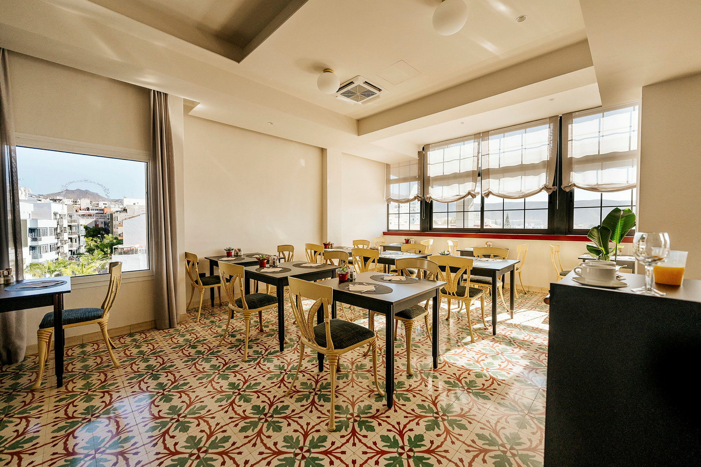 En lys og luftig kafé eller restaurant med bord og stoler, et fargerikt gulvdesign, store vinduer og utsikt over et bylandskap. (AI-generert tekst)