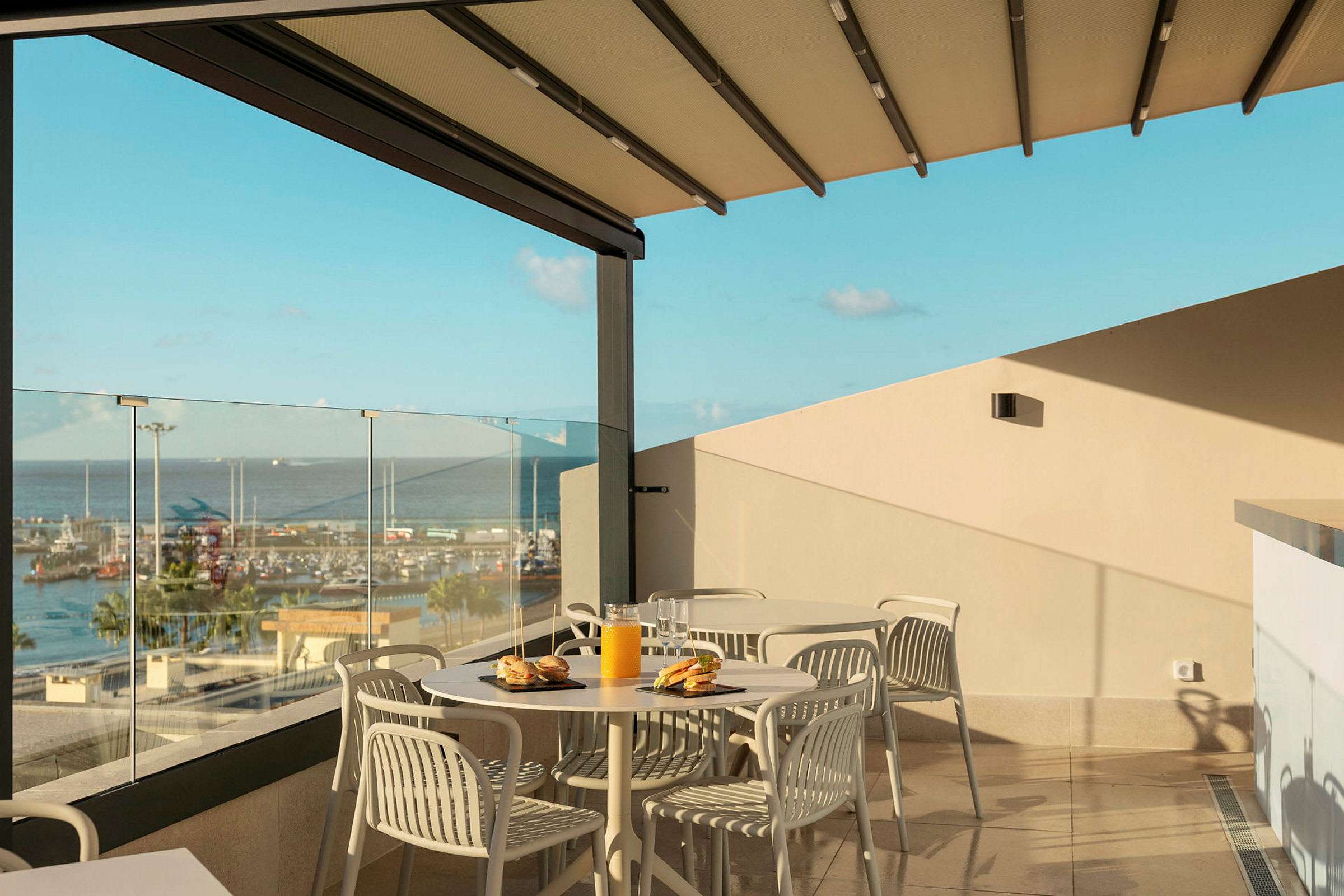 Utsikt fra en terrasse med bord og stoler, glassrekkverk og utsikt over en havn. (AI-generert tekst)
