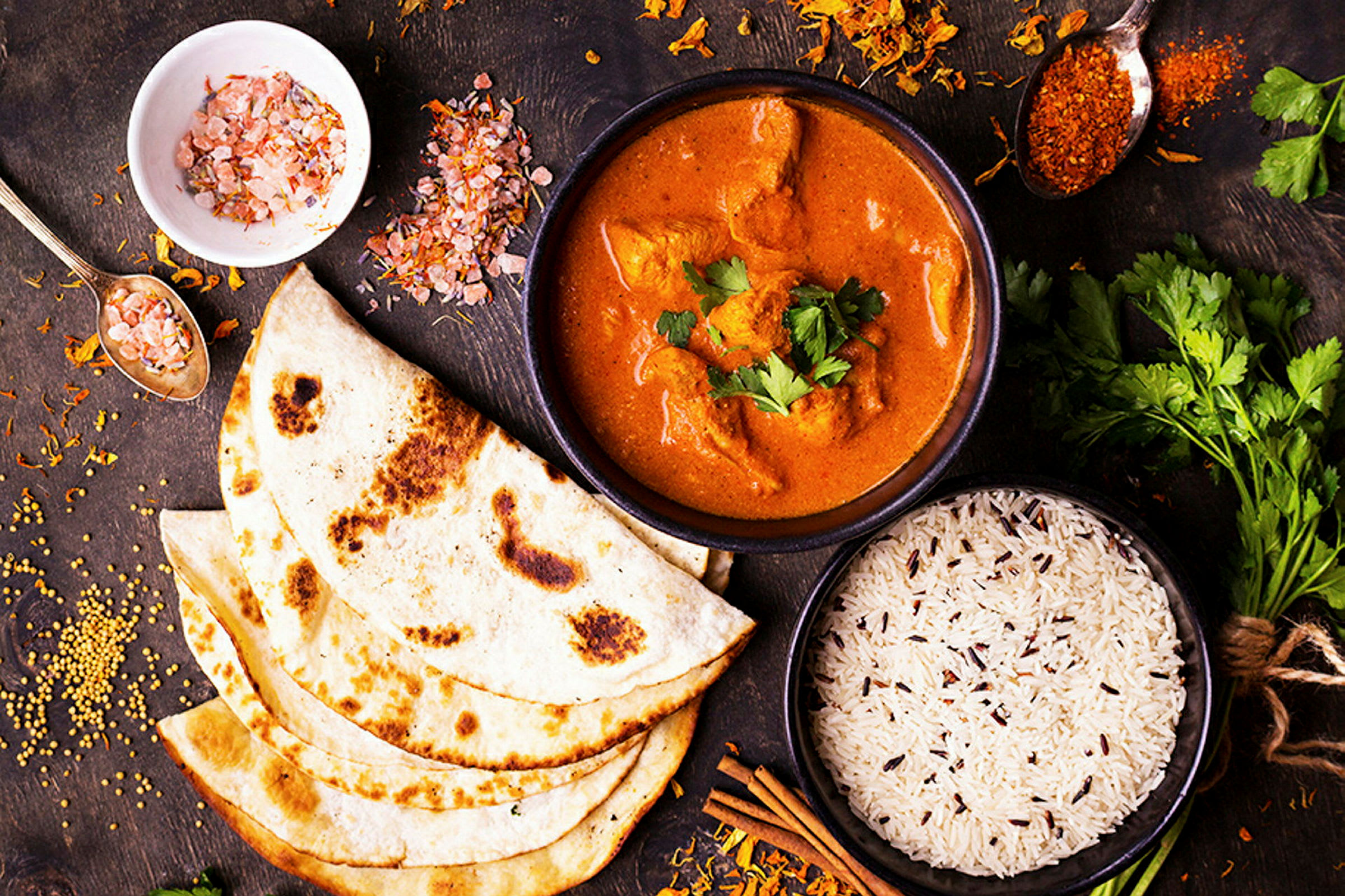 Et oppsett med krydret currygryte, ris og naanbrød, samt krydder og urter på et trebord. (AI-generert tekst)