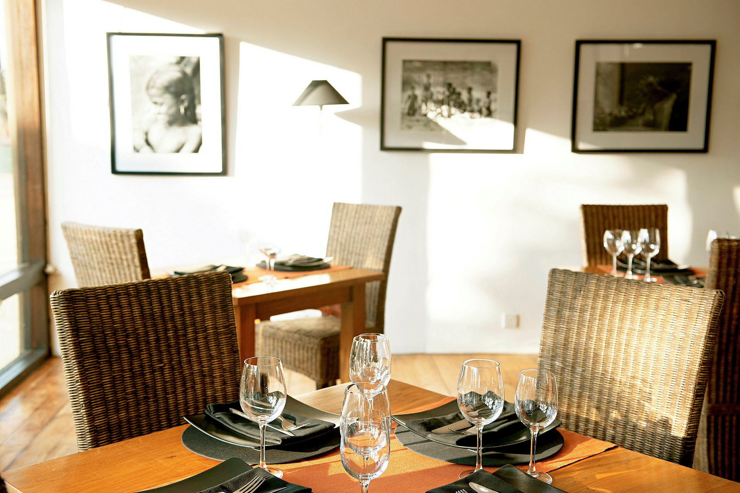 Et elegant restaurantbord med oppdekning, omgitt av rottingstoler og dekorert med glass og servietter. Veggene er pyntet med svart-hvite fotografier. (AI-generert tekst)
