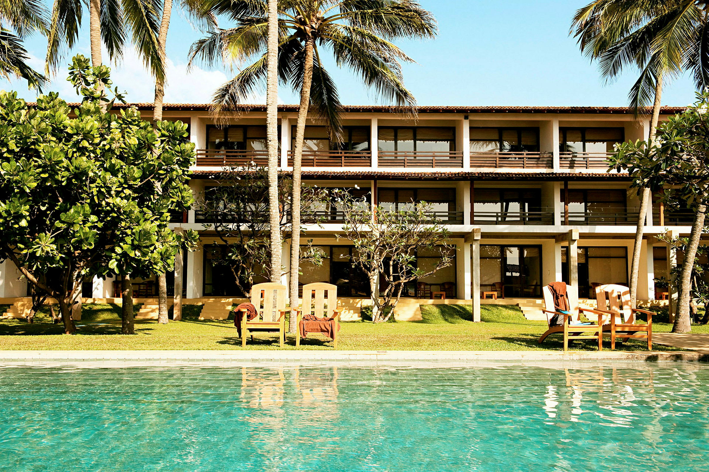 En bygning med flere etasjer og store vinduer kan ses bak et område med trær, plen og stoler. I forgrunnen er det et basseng. (AI-generert tekst)