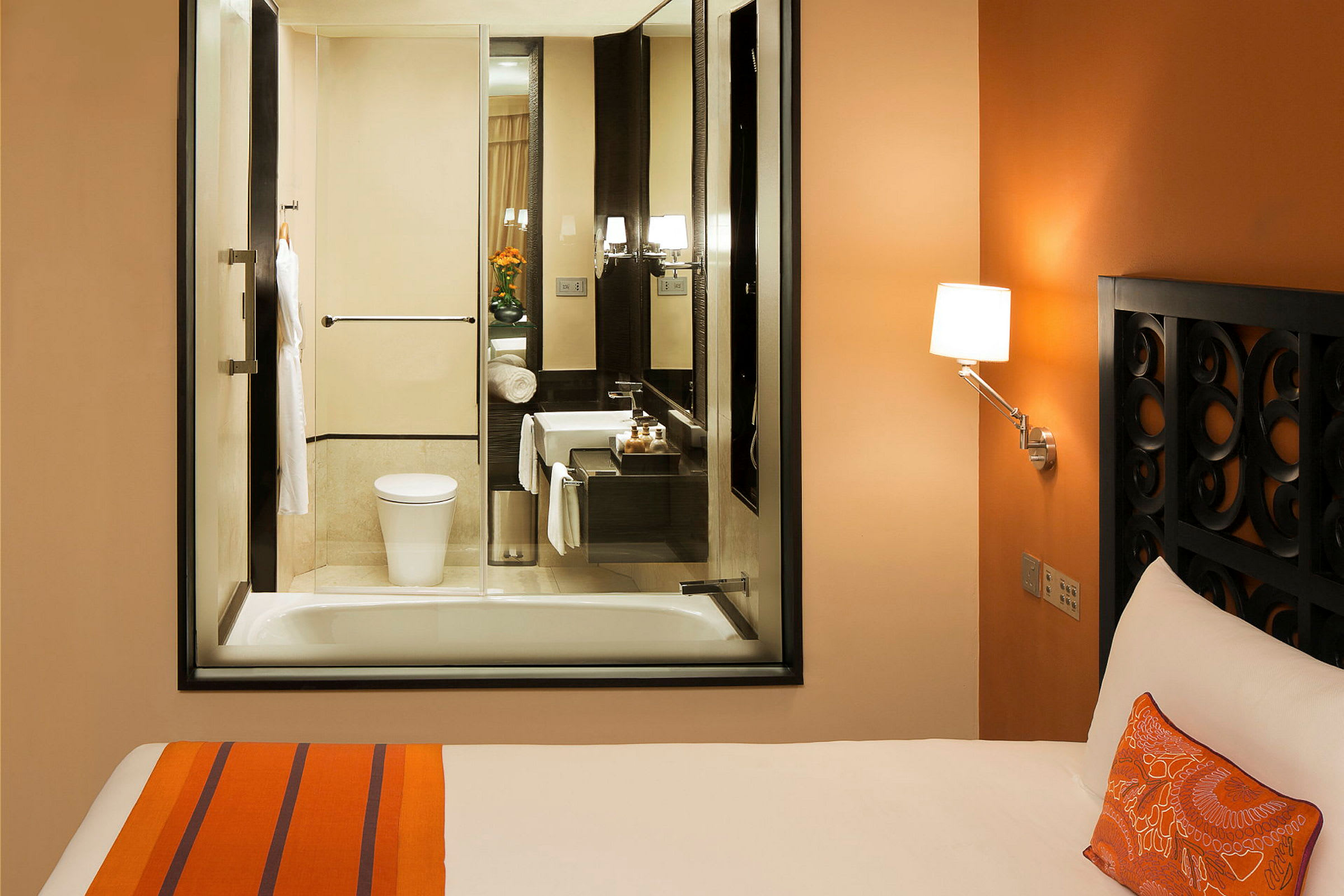 Hotellrom med oransje interiør, seng og et tilstøtende bad med glassvindu. (AI-generert tekst)