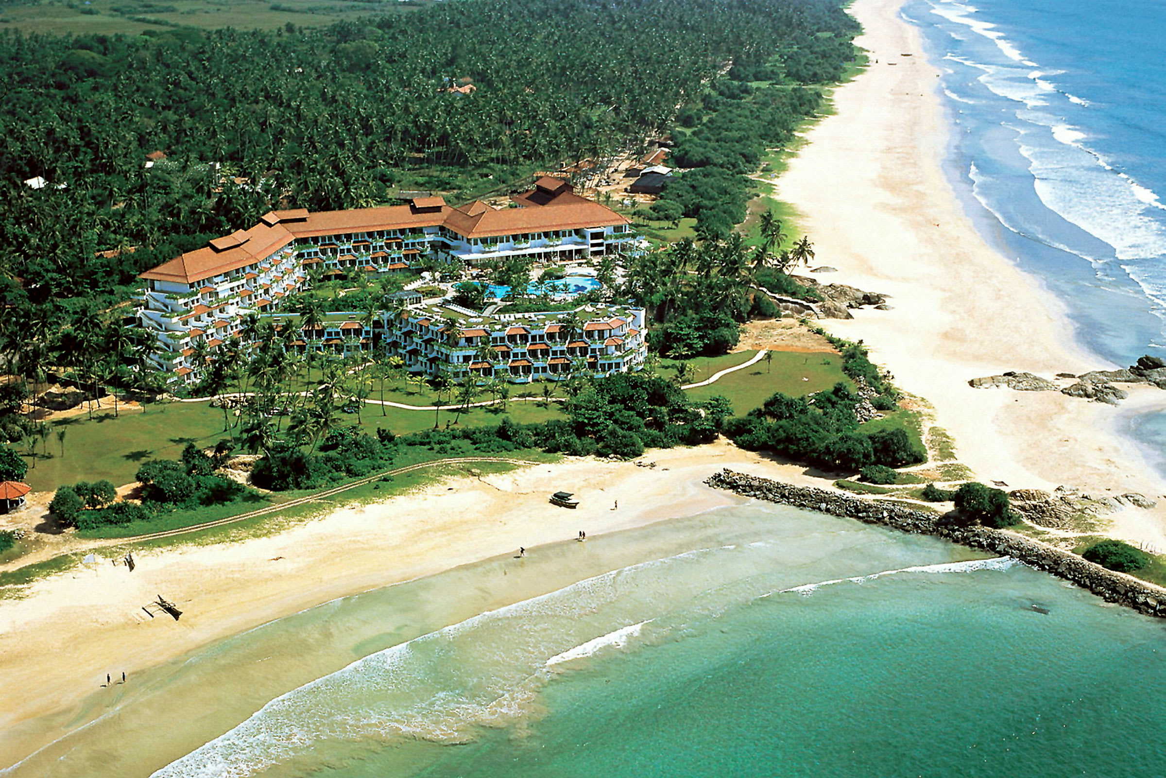 Luftfoto av et hotellkompleks omgitt av grøntområder ved kysten med en lang sandstrand og hav. (AI-generert tekst)