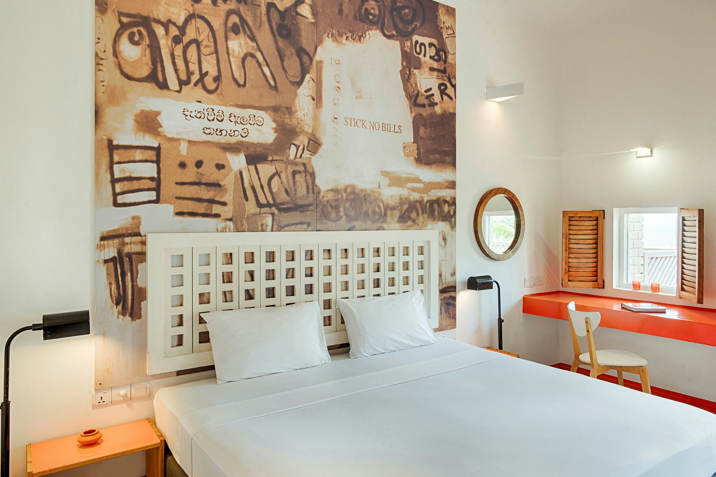 Et hotellrom med en dobbeltseng, et bilde med graffiti-lignende kunst på veggen bak sengen og et skrivebord med speil og stol. (AI-generert tekst)