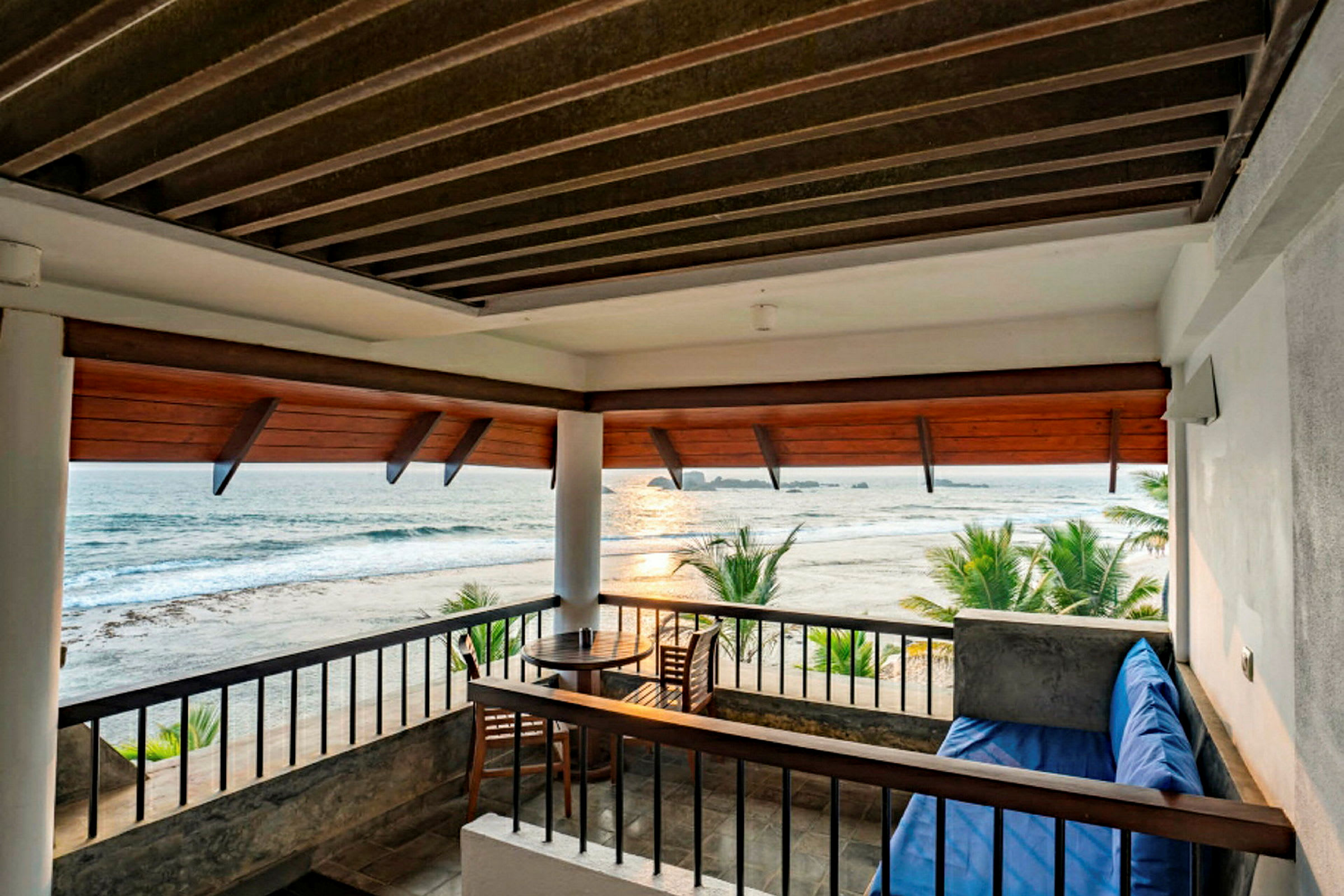 En terrasse med utsikt over stranden og havet. Det er en trestol og et rundt bord samt en sitteplass med en blå pute. (AI-generert tekst)