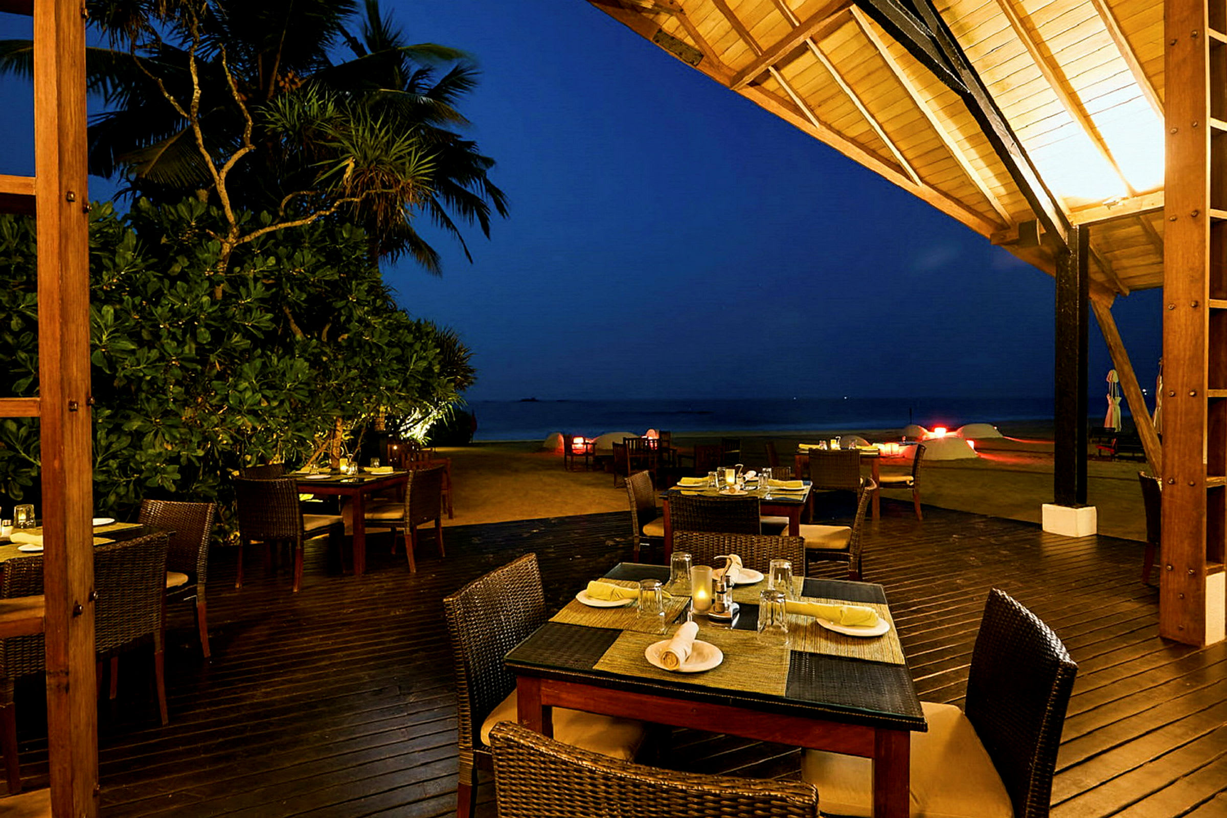 En utendørs restaurant med dekkede bord på en treterrasse med utsikt over stranden og havet. (AI-generert tekst)