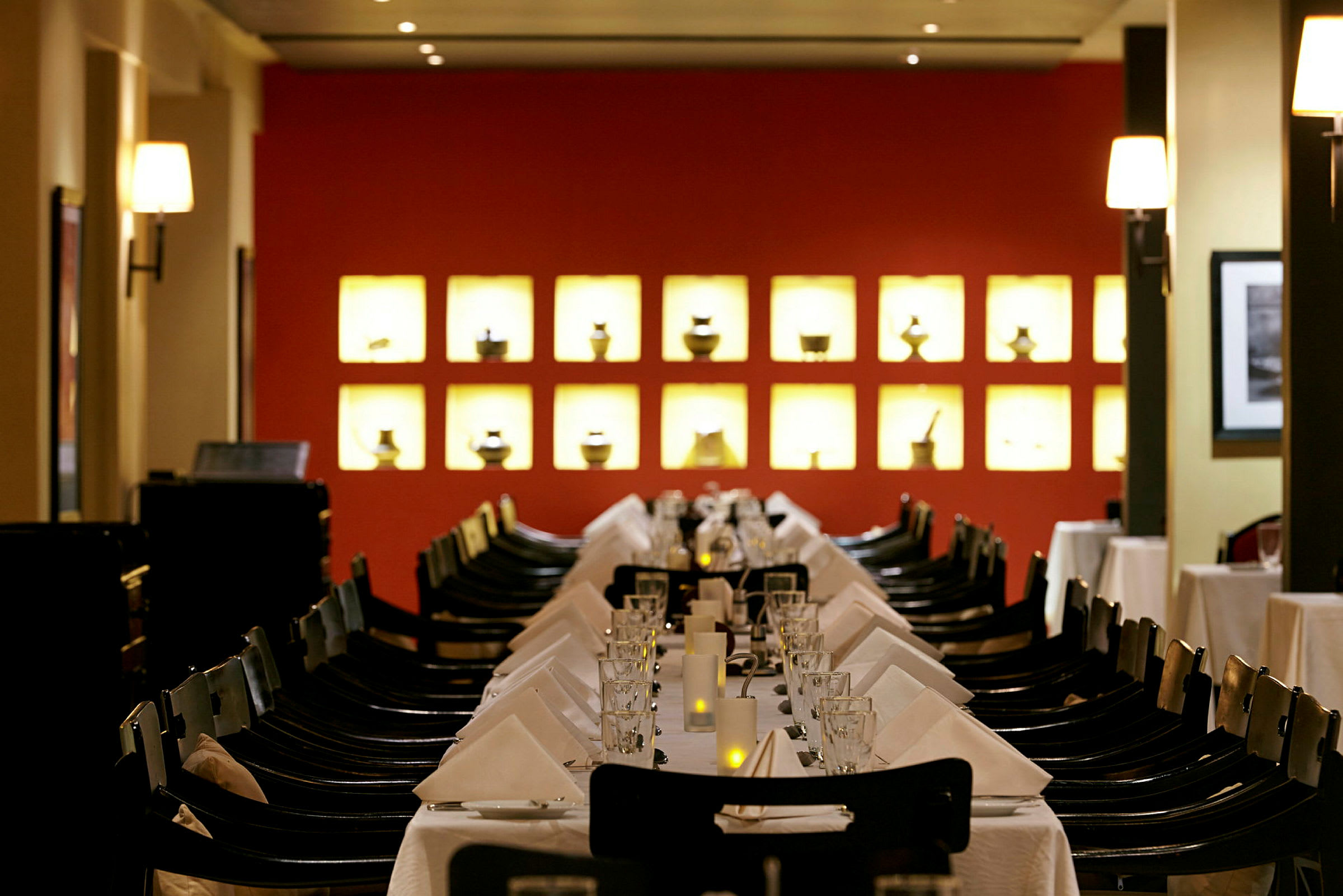 En restaurant med et bord dekket med hvite duker, glass og servietter. I bakgrunnen er det en rød vegg med opplyste små dekorative gjenstander. (AI-generert tekst)