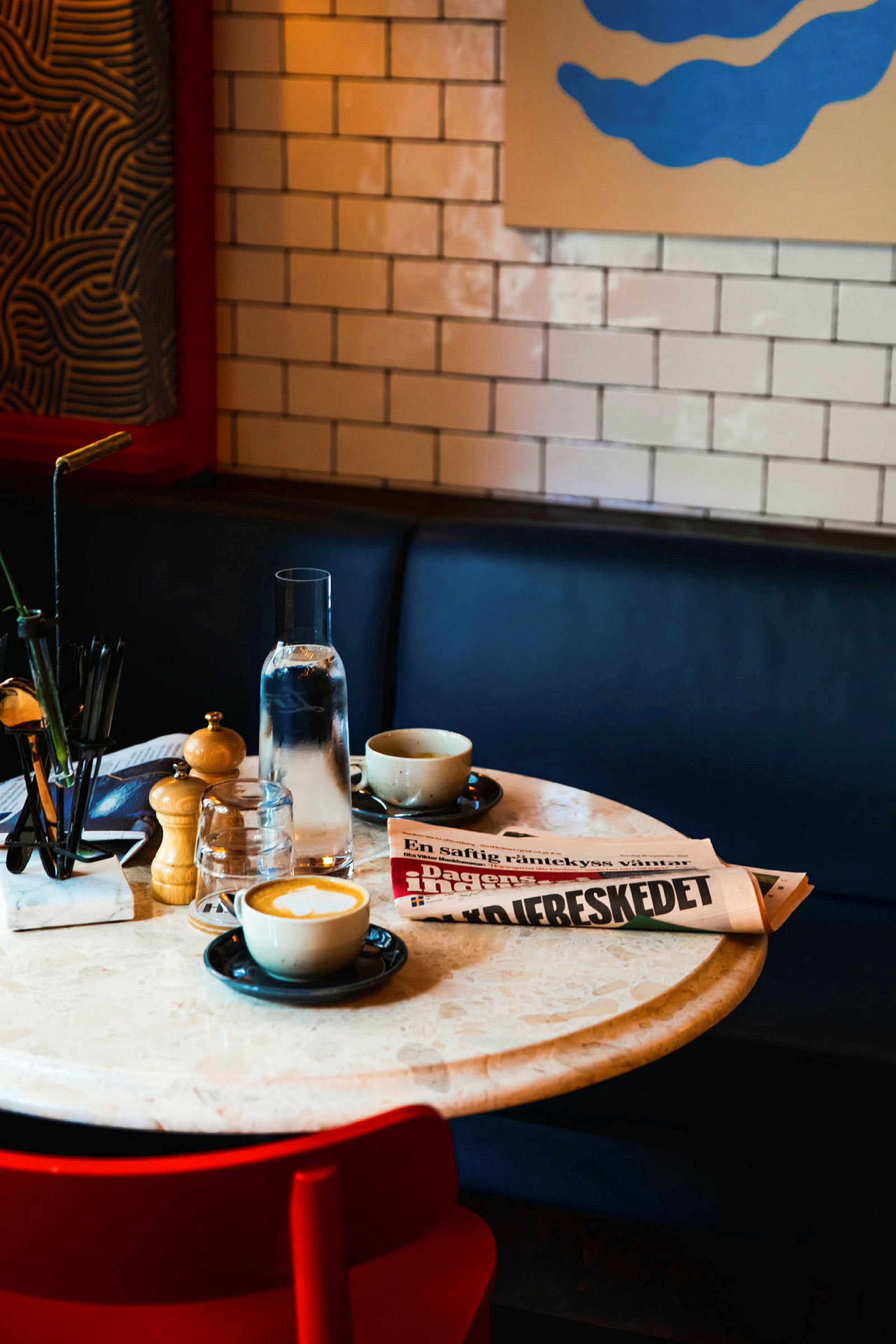 Et kafébord med kaffe, en vannkaraff, et glass og en sammenbrettet avis. (AI-generert tekst)