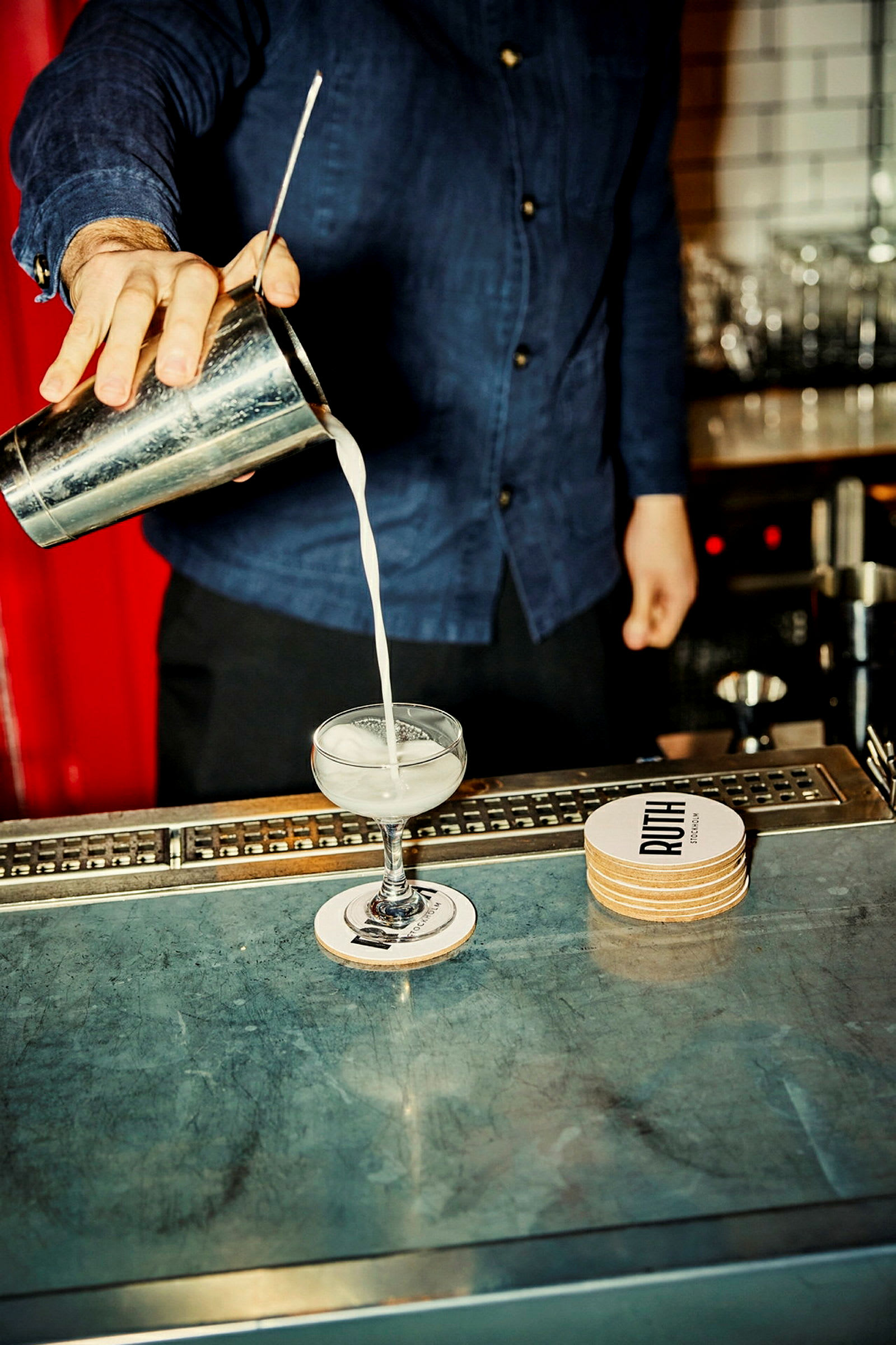 En person heller en drink fra en metallshaker over i et cocktailglass på en bardisk. (AI-generert tekst)