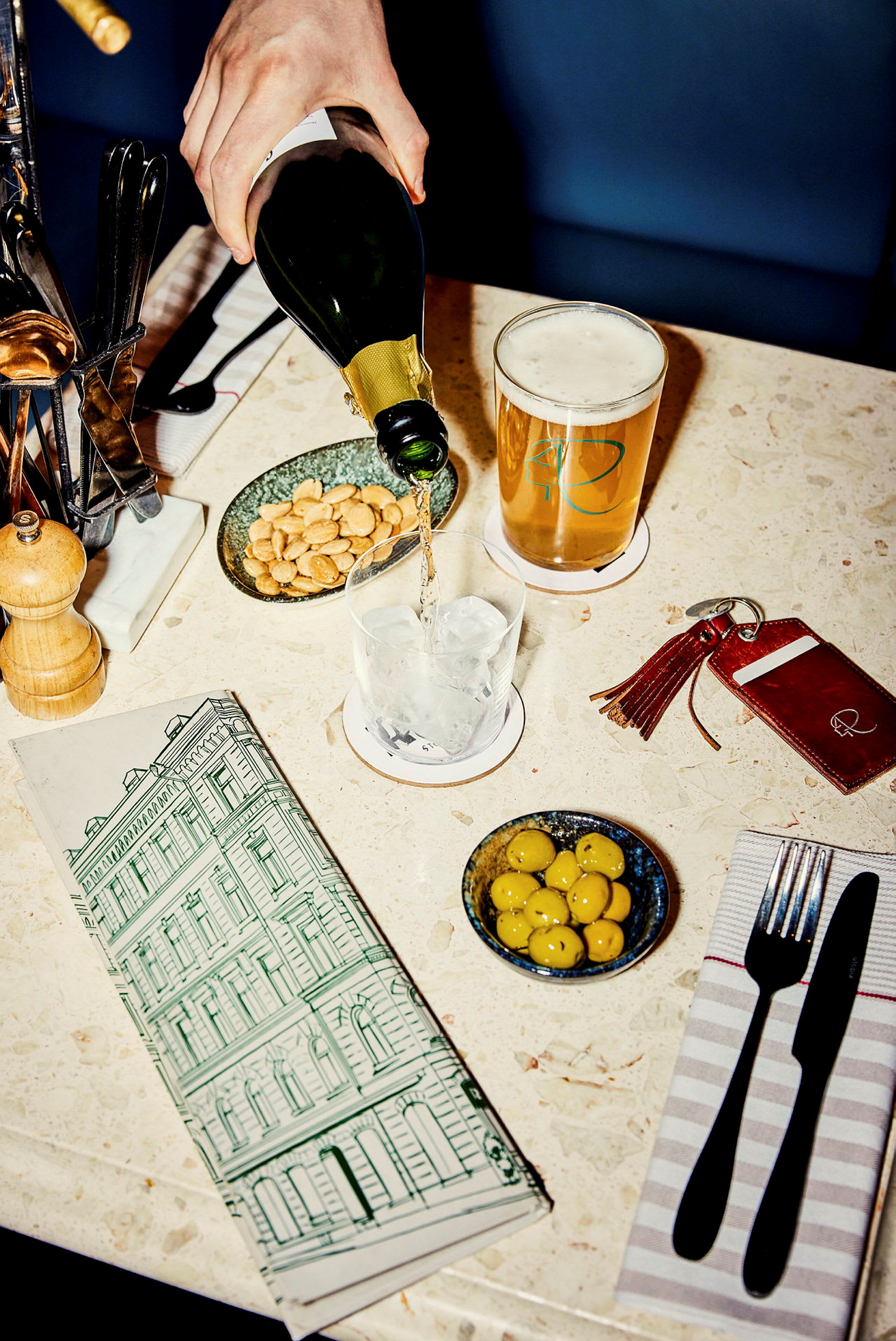 En hånd heller drikke fra en flaske i et glass med is på et dekket bord. På bordet er det også en øl, oliven, nøtter og bestikk. (AI-generert tekst)