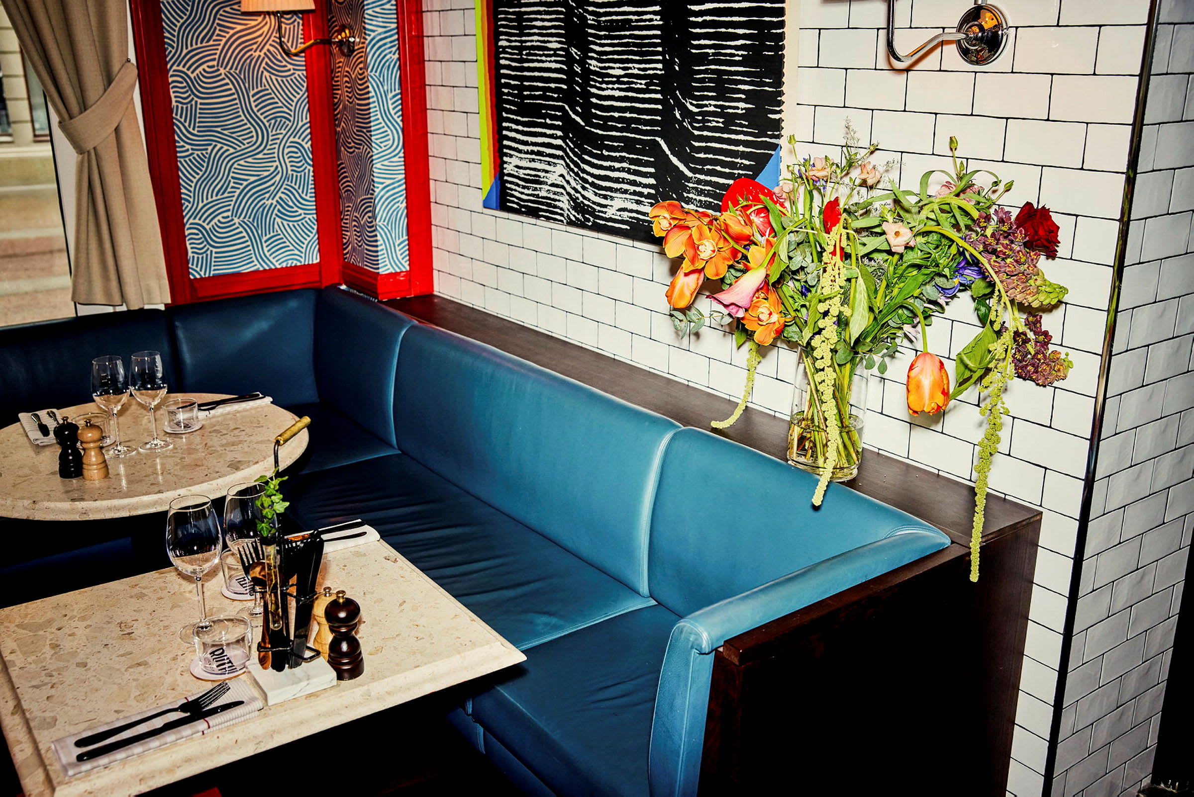 En kafe eller restaurant med et bord og blå sitteplasser. Bordet er dekket med vinglass og krydderkverner. En blomsterbukett står ved siden av på et sidebord. (AI-generert tekst)