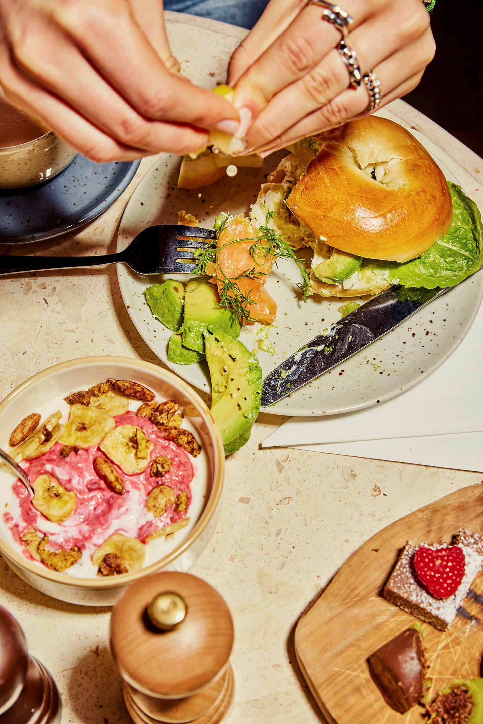 En tallerken med en bagel, avocado og laks, ved siden av en skål med yoghurt og bananchips. (AI-generert tekst)
