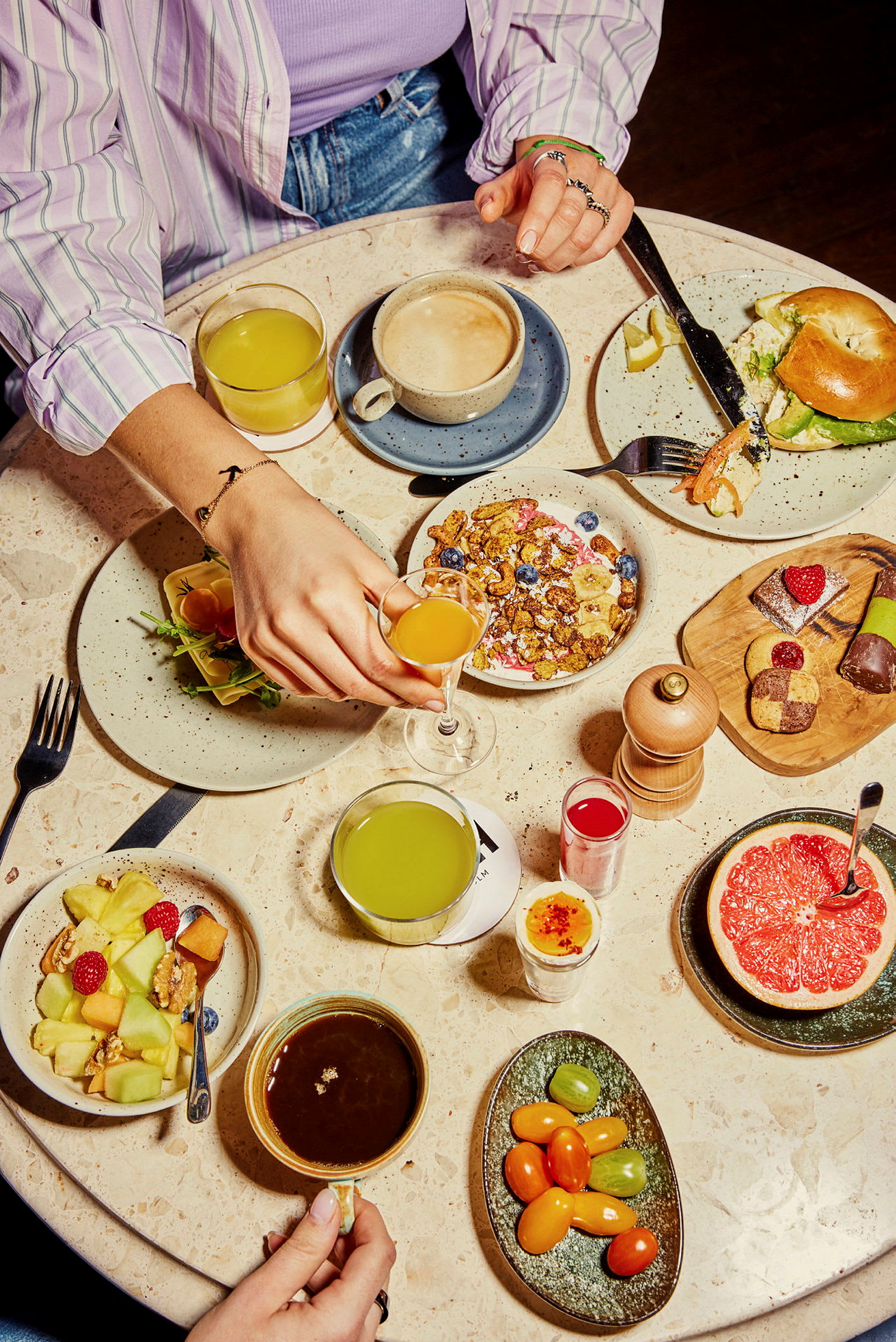 En frokostoppsett med ulike retter, drikke og en hånd som griper et glass. Bordet inneholder blant annet kaffe, juice, salat, grapefrukt og bakverk. (AI-generert tekst)