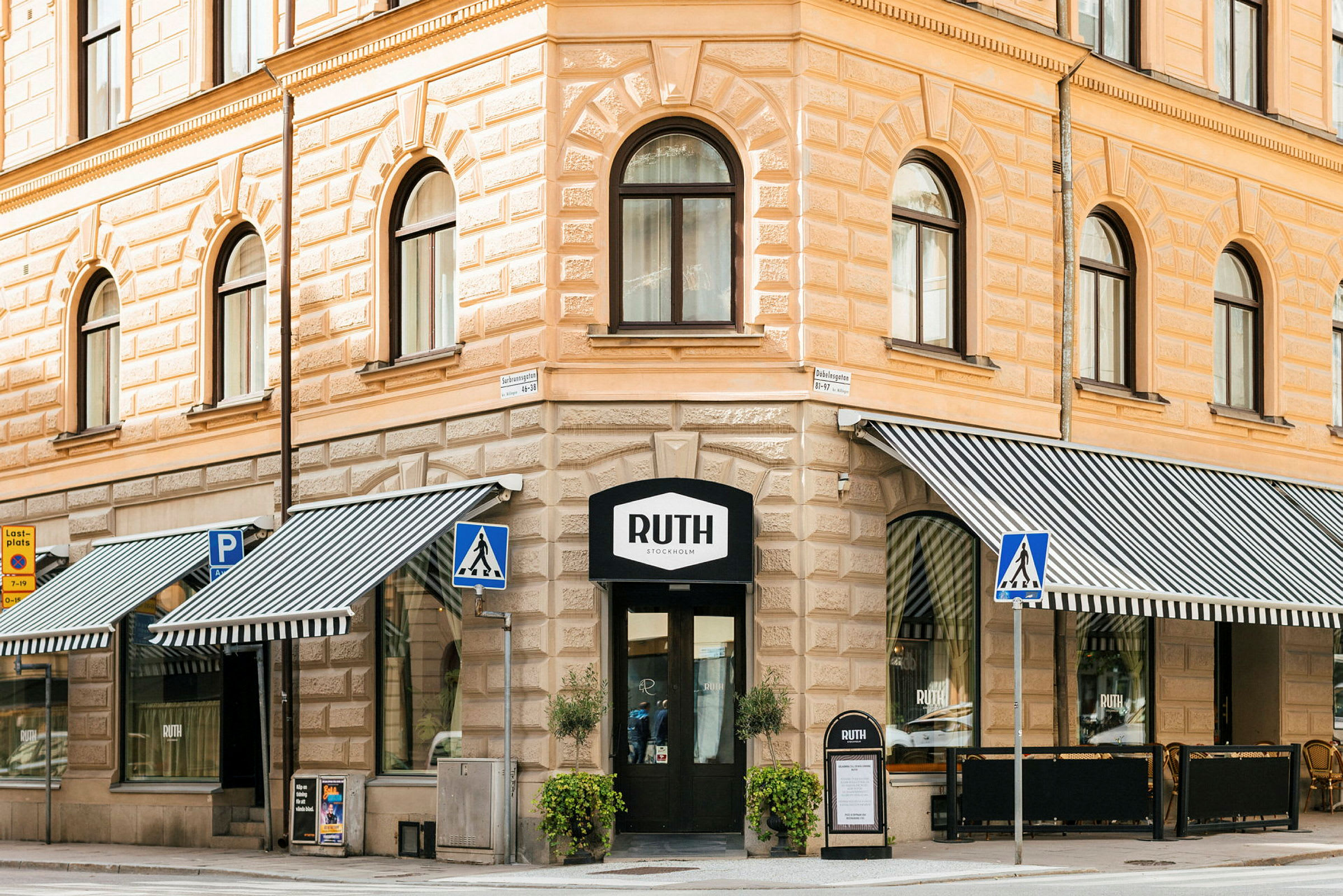 Et hjørnehus i klassisk stil med butikkens skilt Ruth over inngangen. Utstillingsvinduer med stripete markiser, gangvei og stolpeskilt er synlige i forgrunnen. (AI-generert tekst)