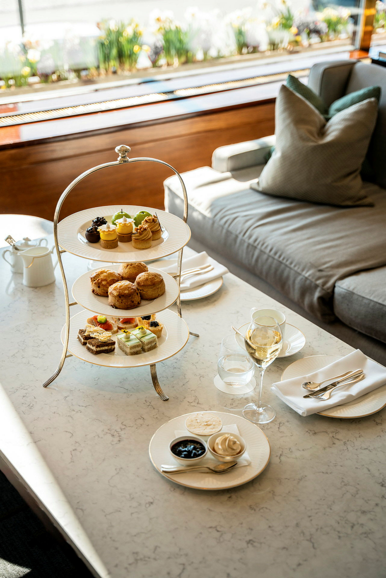 En servering med flere nivåer med kaker, scones og smørbrød står på et bord ved en sofa. (AI-generert tekst)