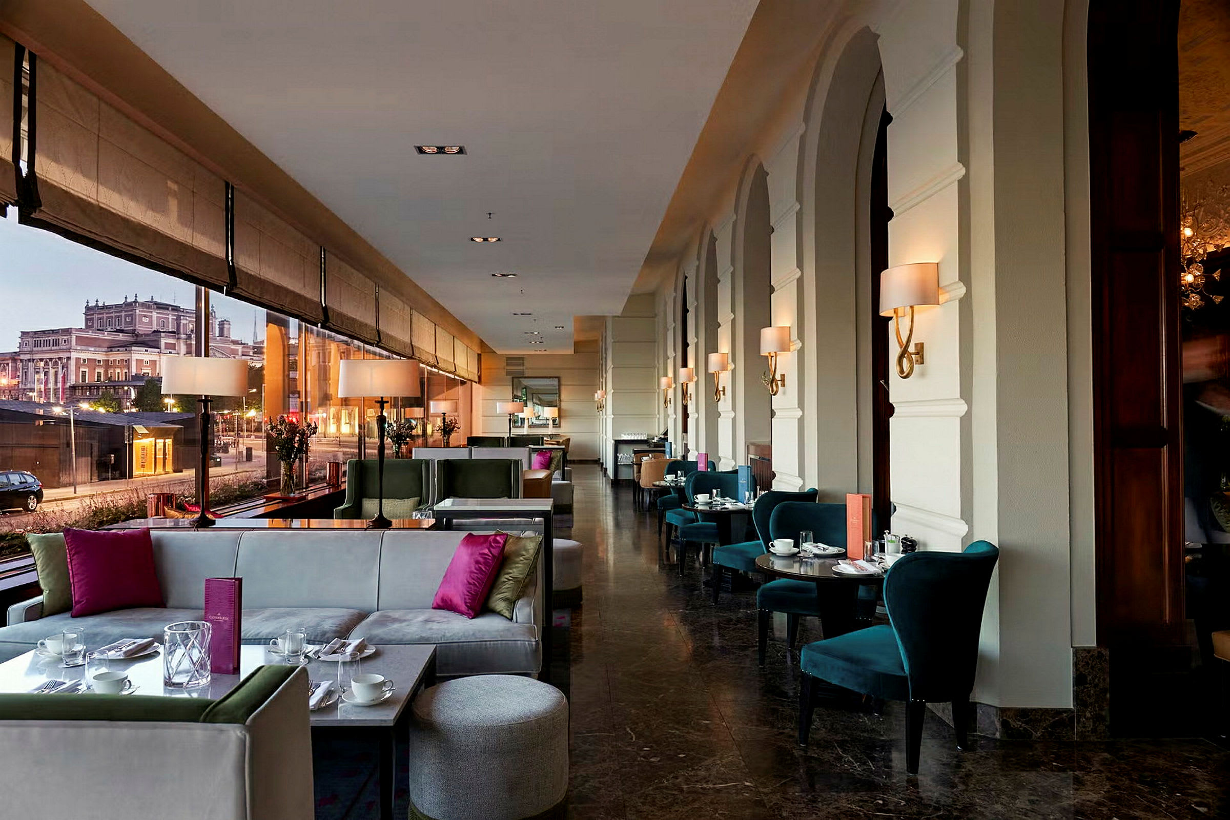 Et elegant og moderne restaurant- eller kaféområde med store vinduer og utsikt over en historisk bygning utenfor. Møblene består av komfortable sofaer og stoler. (AI-generert tekst)