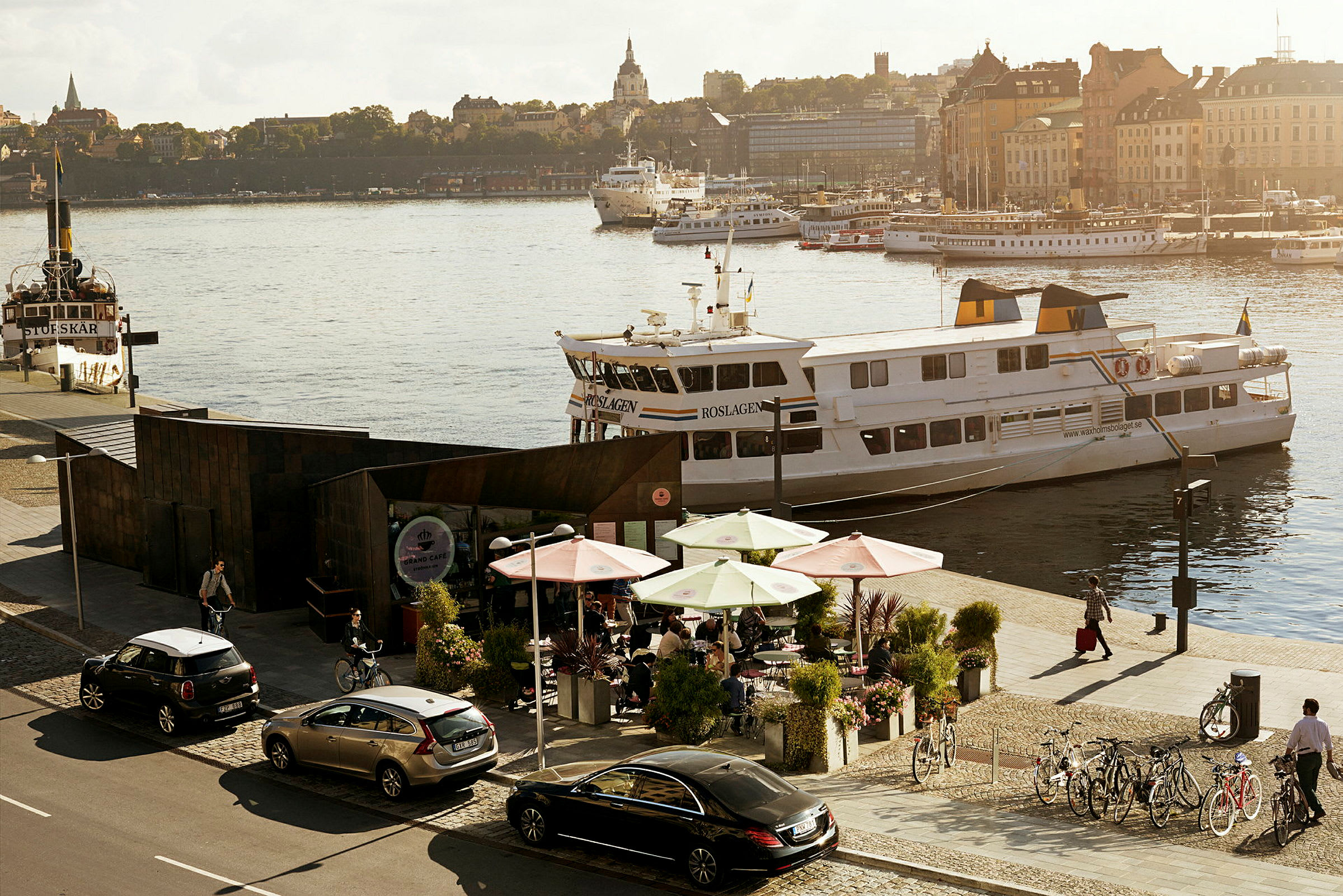 Bilde av en by ved vannet med kafébord på bryggen, båter og et livlig miljø. (AI-generert tekst)