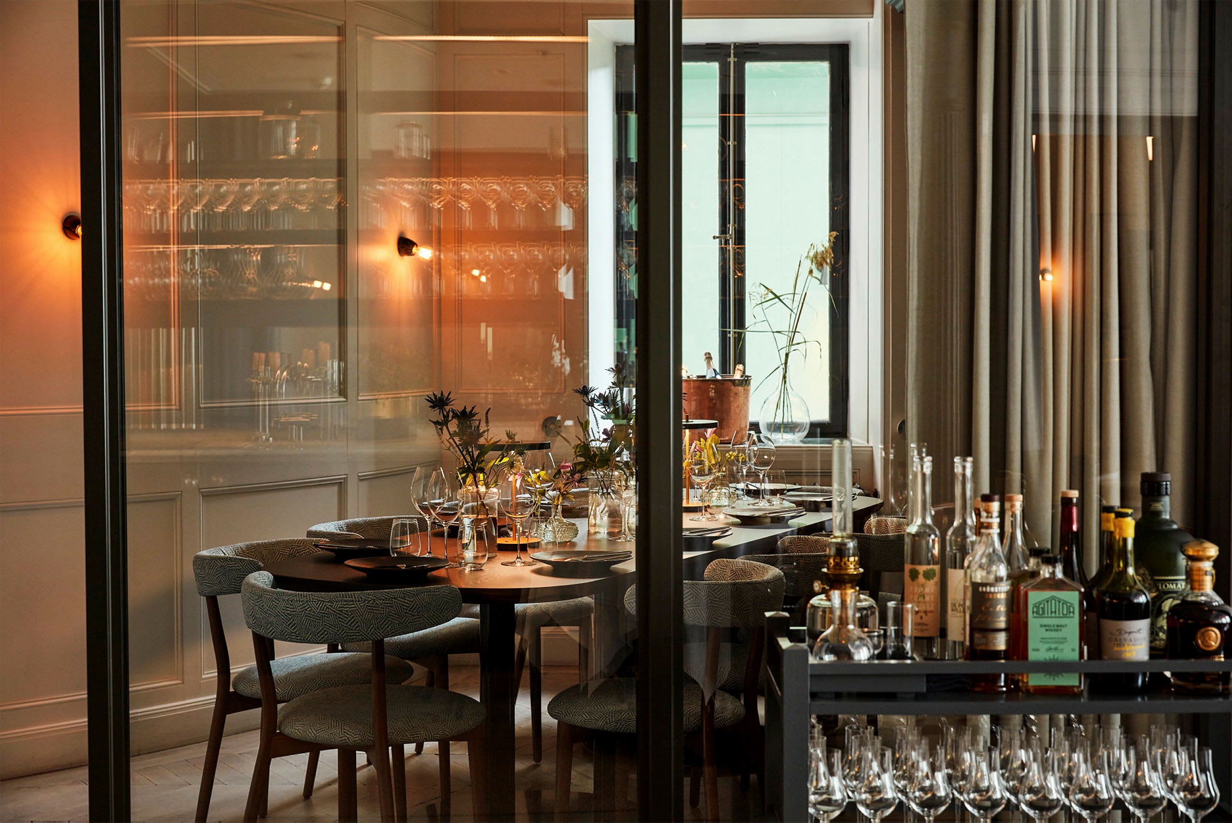 Bildet viser et elegant restaurantområde med et dekket bord dekorert med glass og blomster. En glassvegg skimtes i forgrunnen, og en vogn med drikkevarer og glass står nær. (AI-generert tekst)