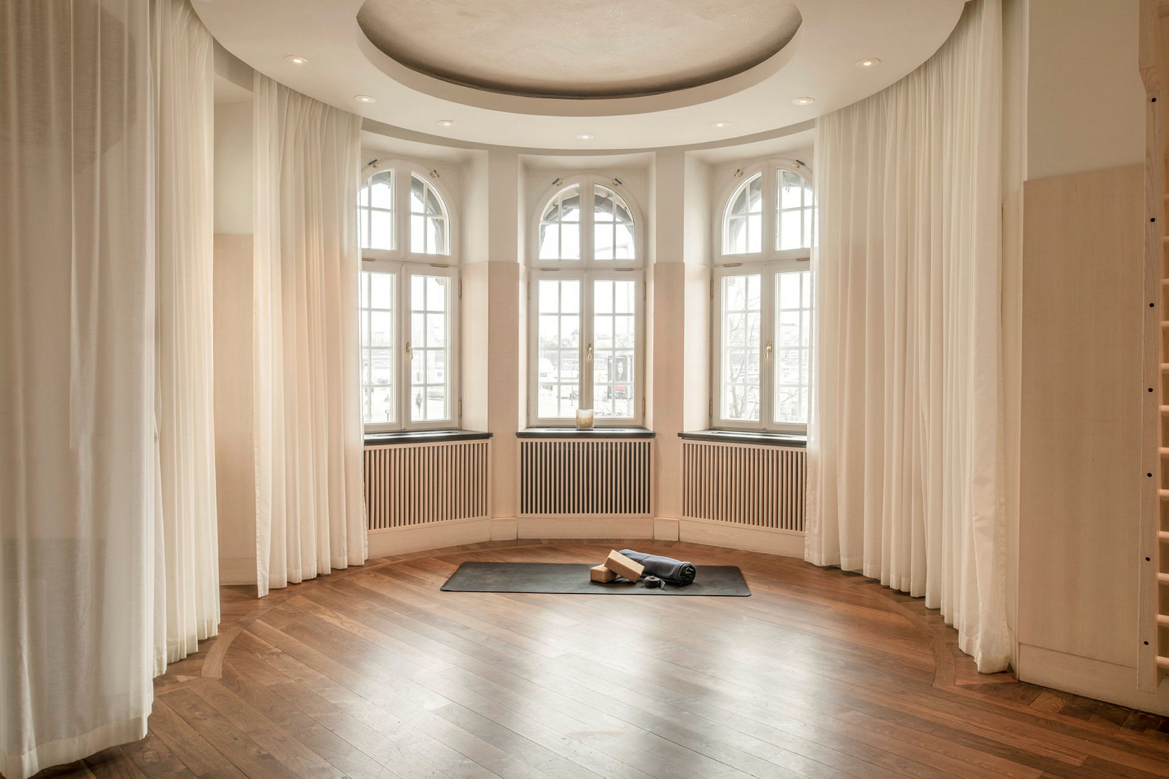 Et rom med buede vinduer og lyse gardiner. På gulvet ligger en treningsmatte sammen med yoga-utstyr. (AI-generert tekst)