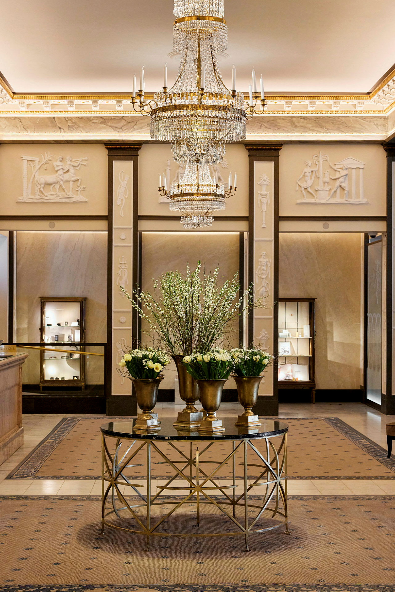 En luksuriøs lobby med en stor lysekrone, kunstnerisk dekorerte vegger og et bord med gullfargede blomsterurner fylt med hvite og grønne blomster. (AI-generert tekst)