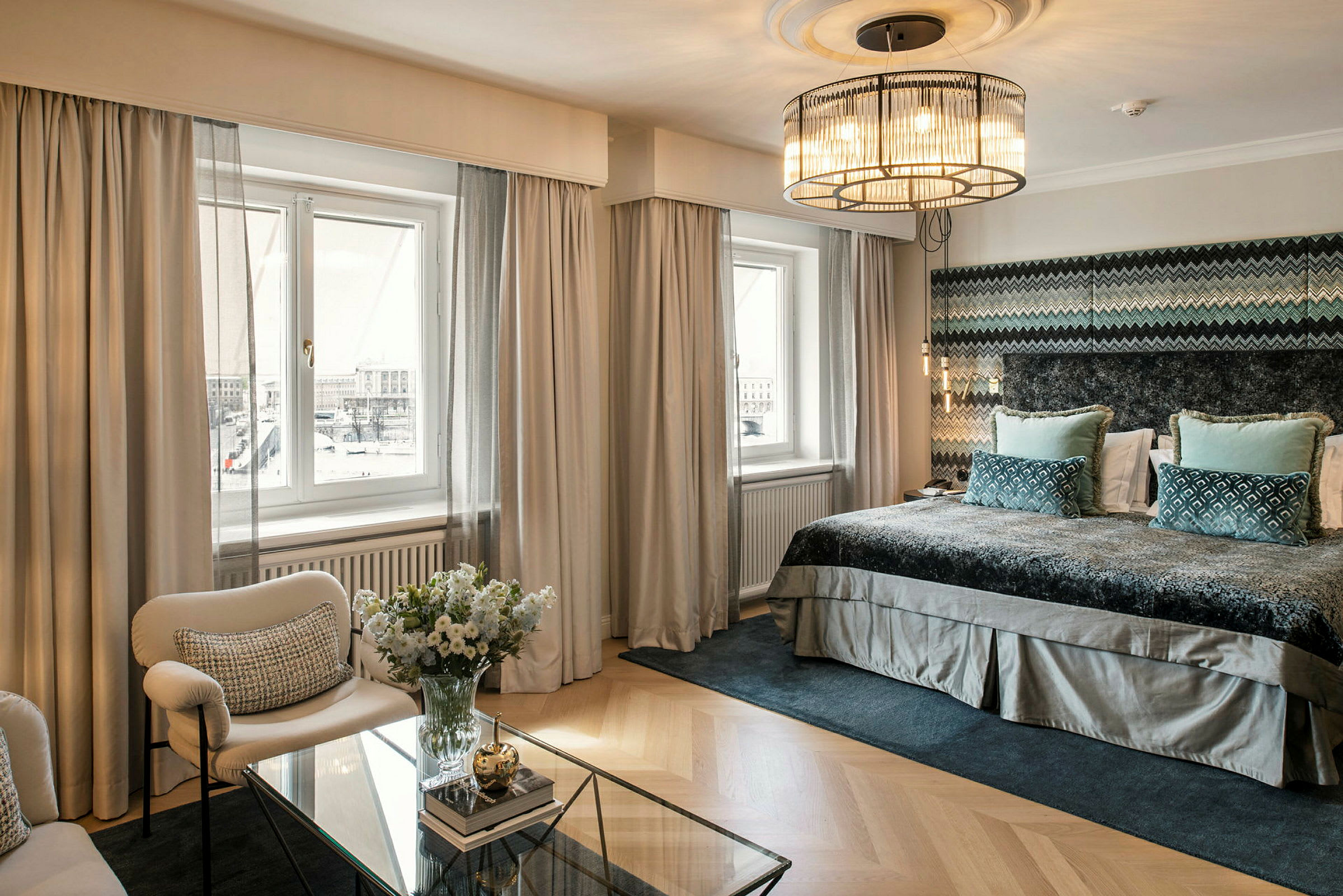 Et elegant hotellrom med store vinduer, bred seng, komfortable sittemøbler og et glassbord med dekorativ oppsett. (AI-generert tekst)