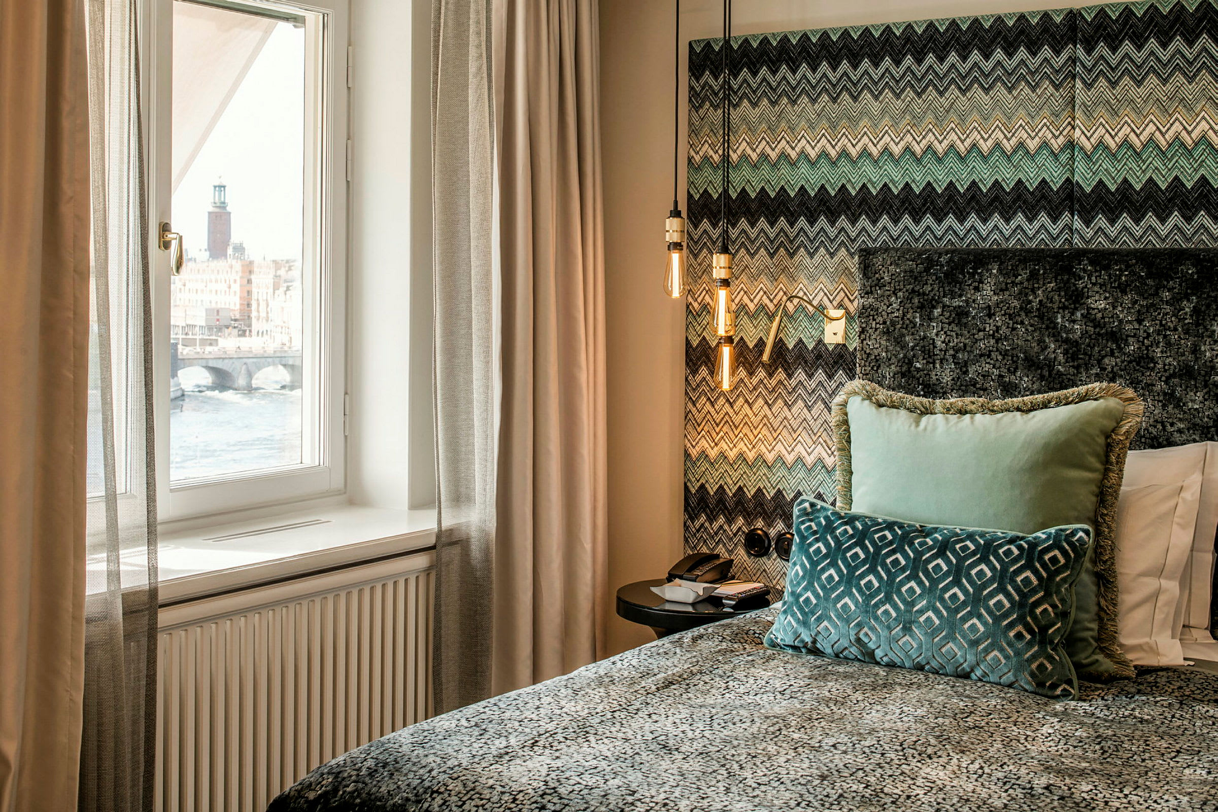 Et moderne hotellrom med et vindu som viser utsikten over vannet og rådhuset. Sengen er dekorert med fargerike puter og et mønstret overkast. (AI-generert tekst)