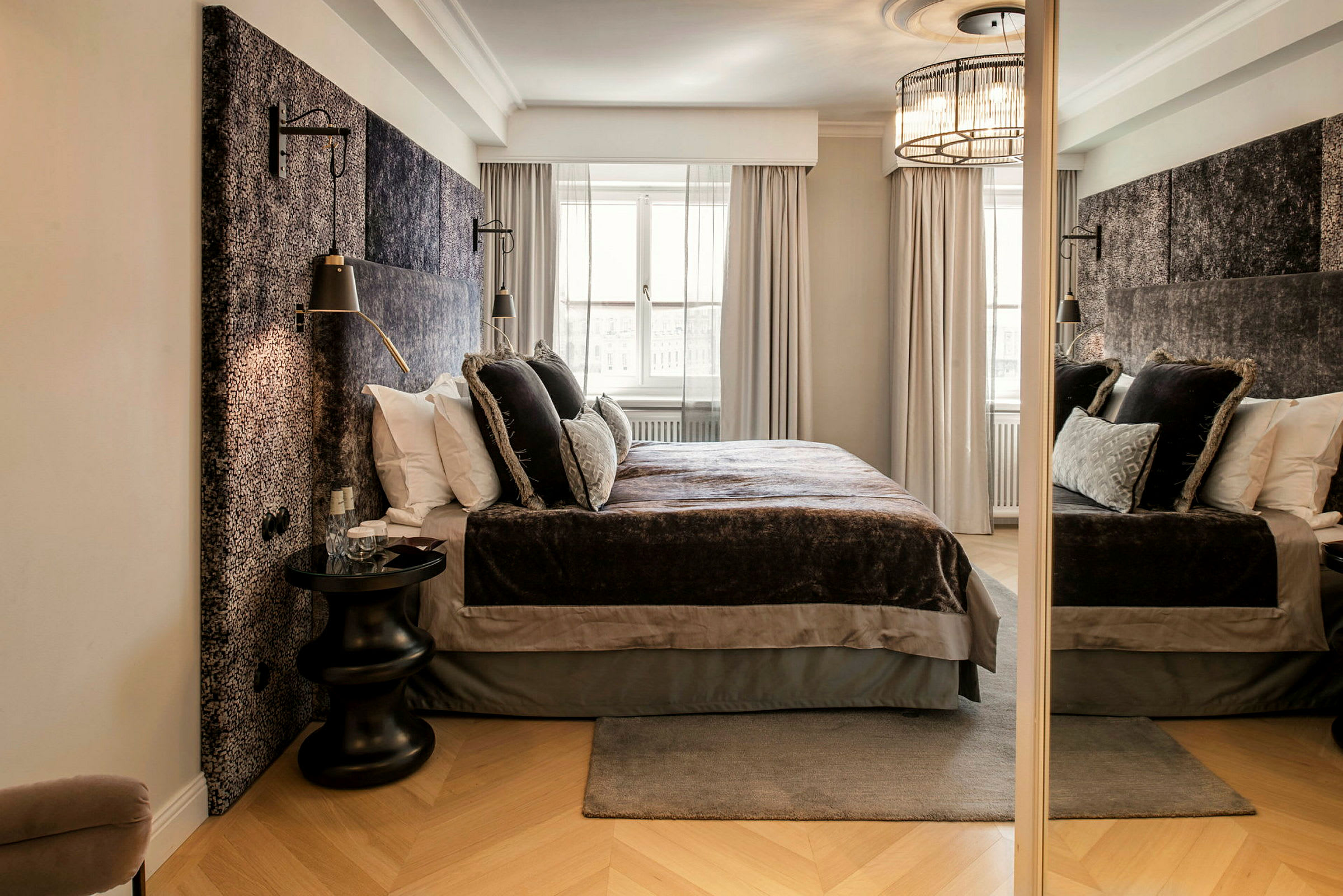 Et elegant hotellrom med en stor seng pyntet med myke sengetepper og dekorative puter. Rommet har lyse gardiner ved vinduet og et speil på siden. (AI-generert tekst)