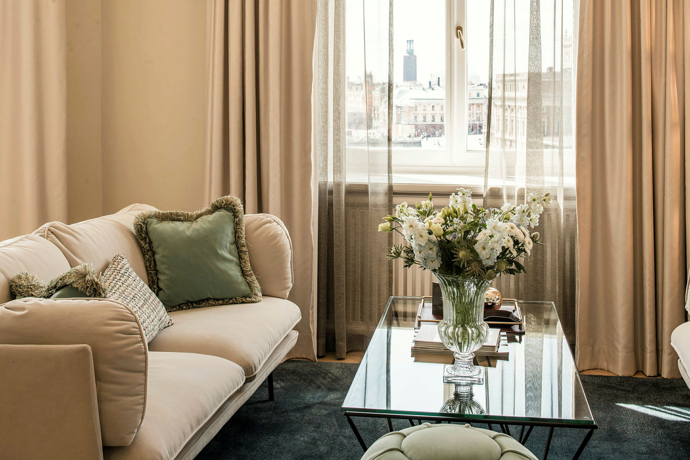 En stue med en lys sofa, puter med farger, et glassbord med en blomster vase og gardiner, samt utsikt mot et tårn og bygninger utenfor vinduet. (AI-generert tekst)