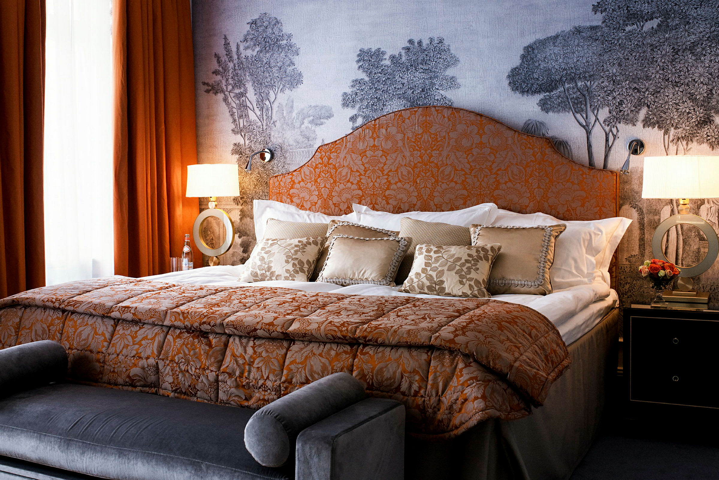 Et elegant hotellrom med en stor seng, dekorert med orange og hvite sengetepper samt flere puter. Bak sengen er en vegg med motiv av trær. (AI-generert tekst)