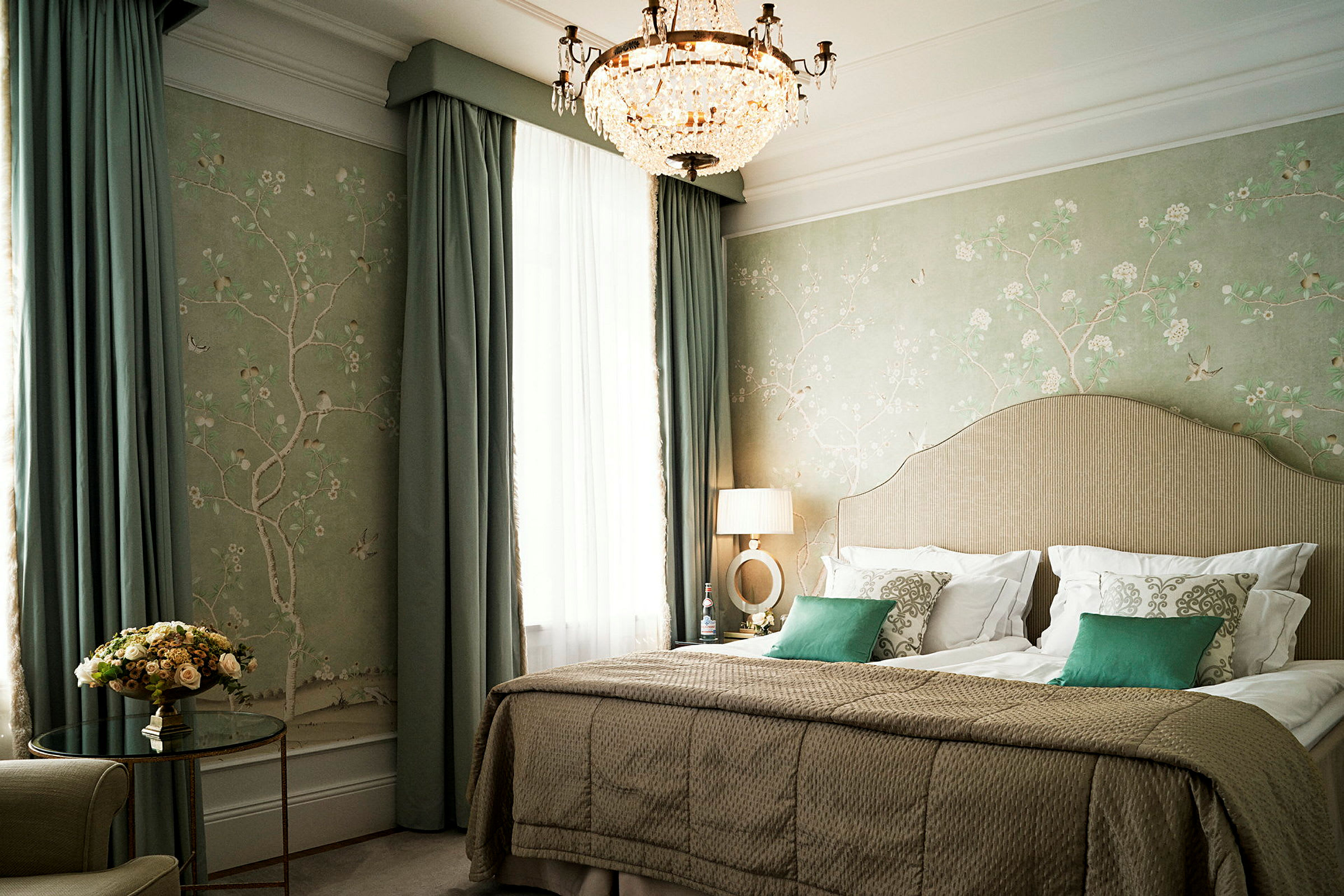Et elegant hotellrom med en stor seng, dekorative puter, grønne gardiner og en lysekrone i taket. (AI-generert tekst)
