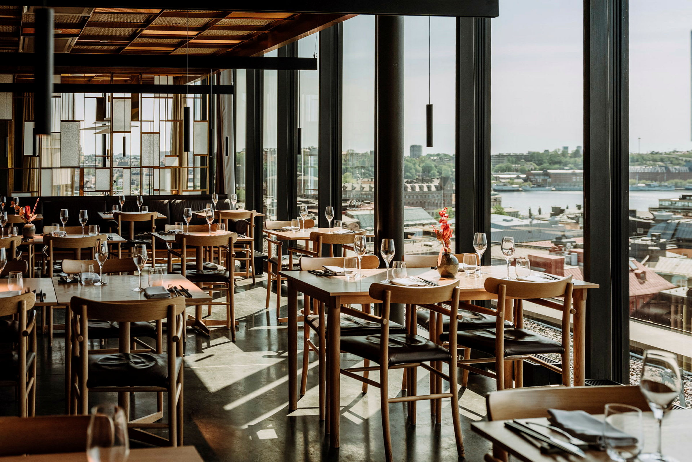 En stilfull restaurant med trebord og stoler, glass som reflekterer lys, og panoramautsikt over en by. (AI-generert tekst)