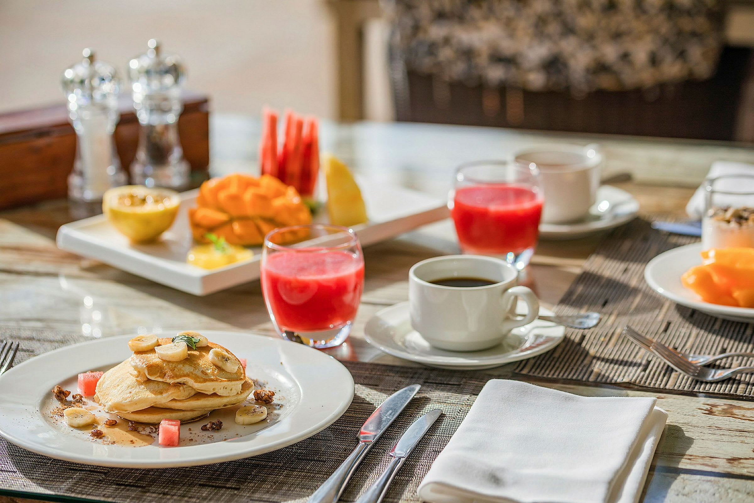 En frokostoppsett med pannekaker, frukt, juice og kaffe på et bord. (AI-generert tekst)