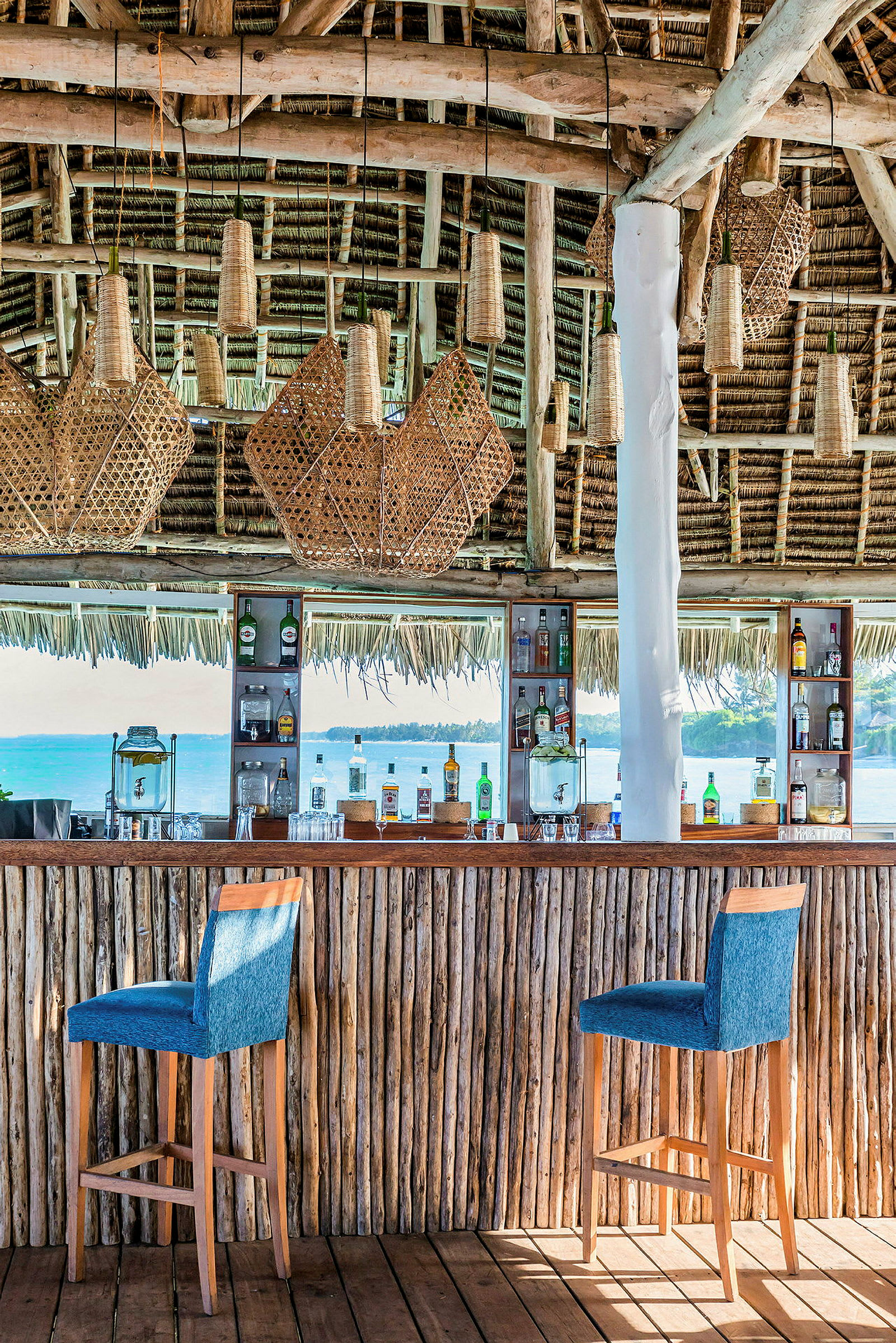 En strandbar med trestamme og halmtak. Flere flettede lamper henger fra taket, og baren har to høye stoler samt forskjellige flasker med drikke på hyllene. Utsikten viser blått hav og en kyst i bakgrunnen. (AI-generert tekst)