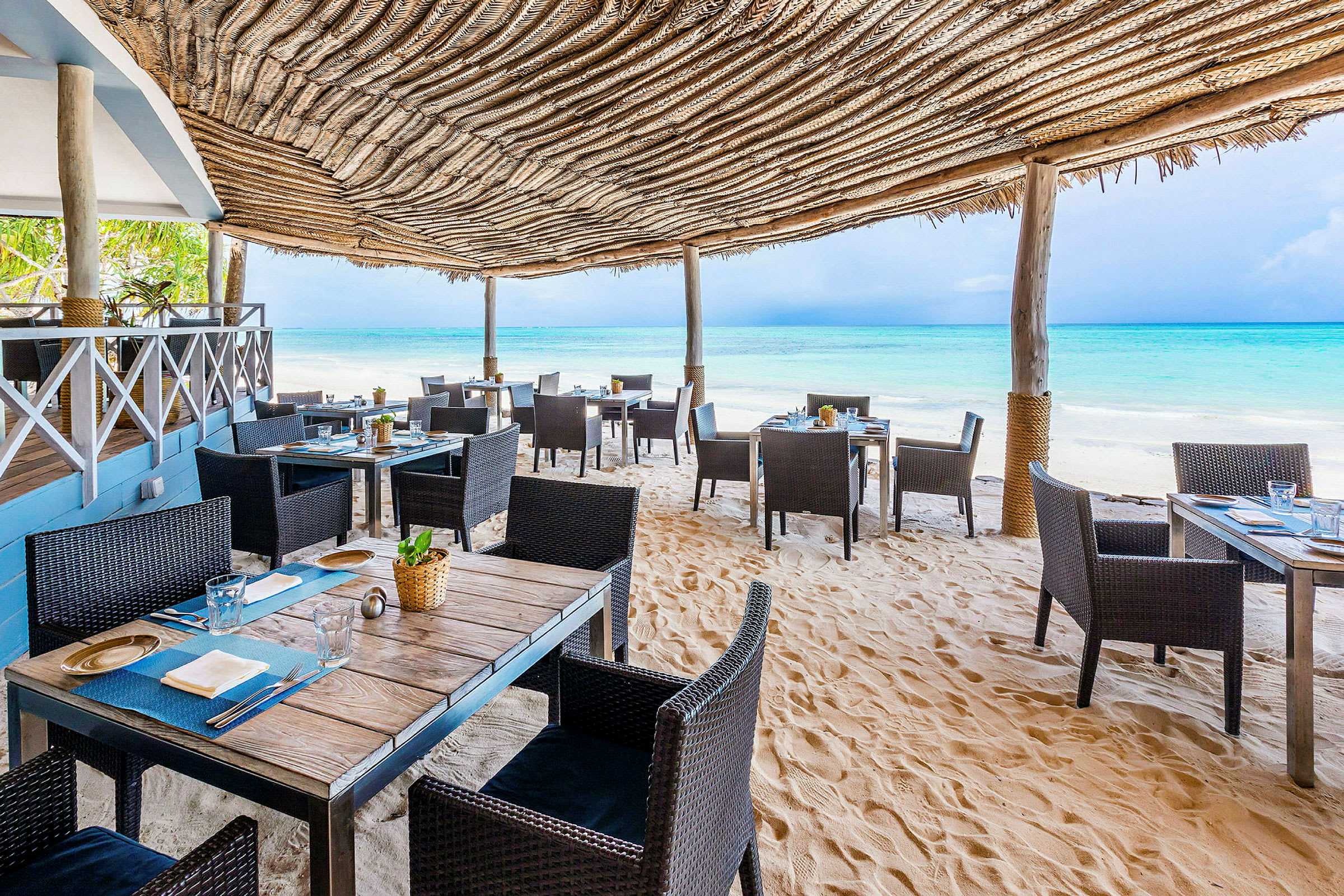 En strandrestaurant med bord og stoler på sanden under et tak av naturmaterialer, med utsikt over det blå havet. (AI-generert tekst)