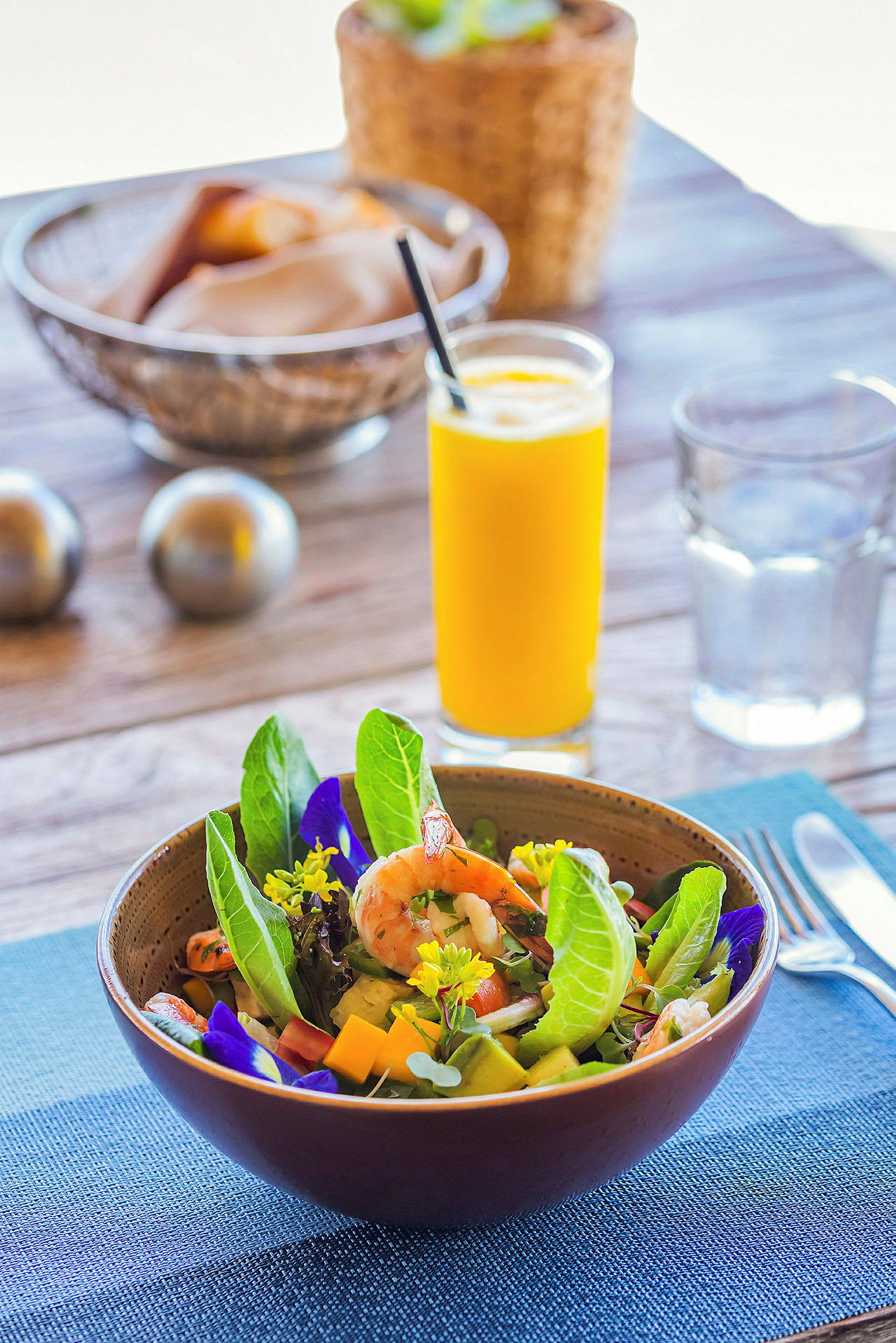 En salat i en bolle med reker, grønne blader og blomster. Et glass appelsinjuice og brød i bakgrunnen. (AI-generert tekst)