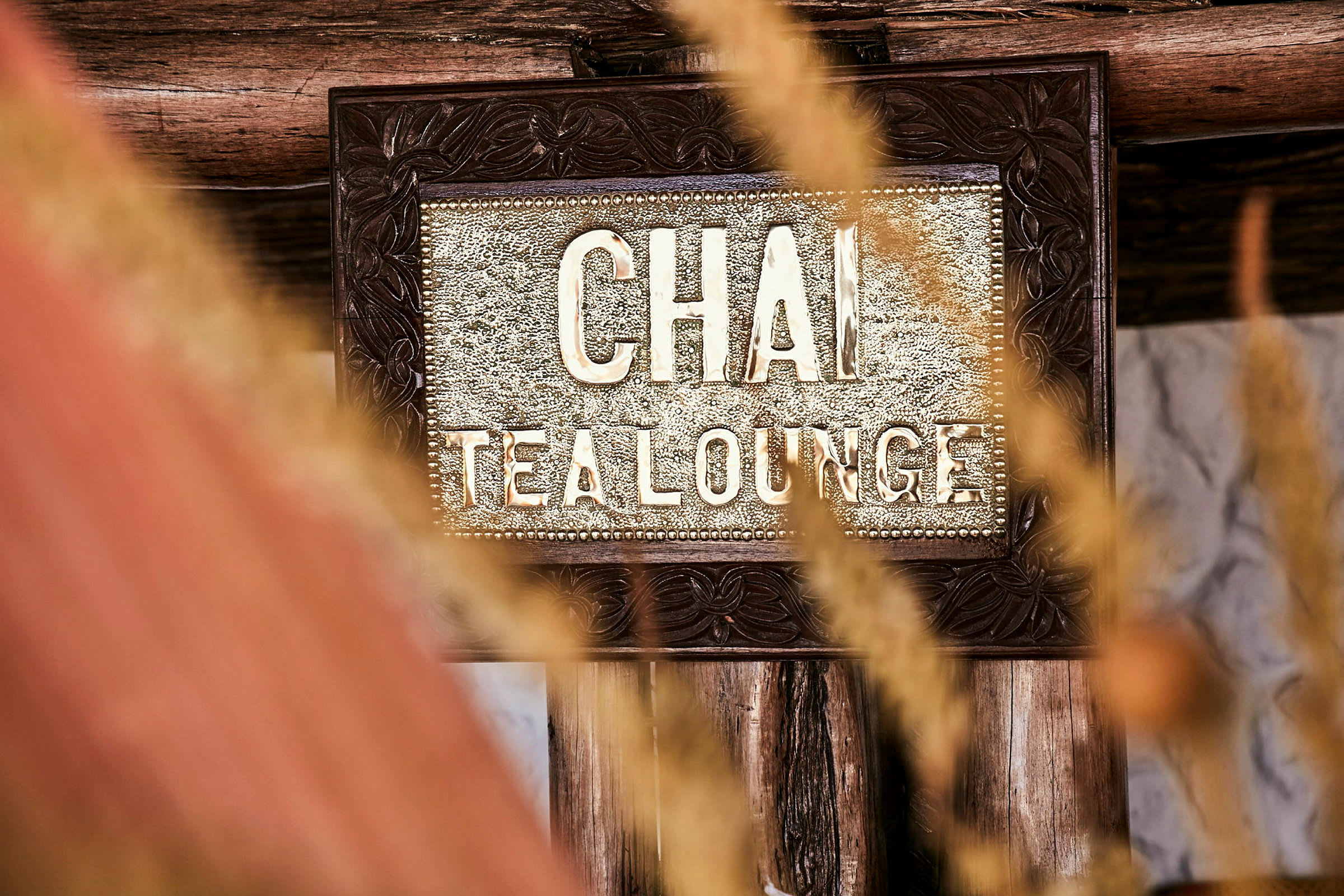 Et nærbilde av et skilt hvor det står 'Chai Tea Lounge', omgitt av tredekorasjoner og visnende gress i forgrunnen. (AI-generert tekst)