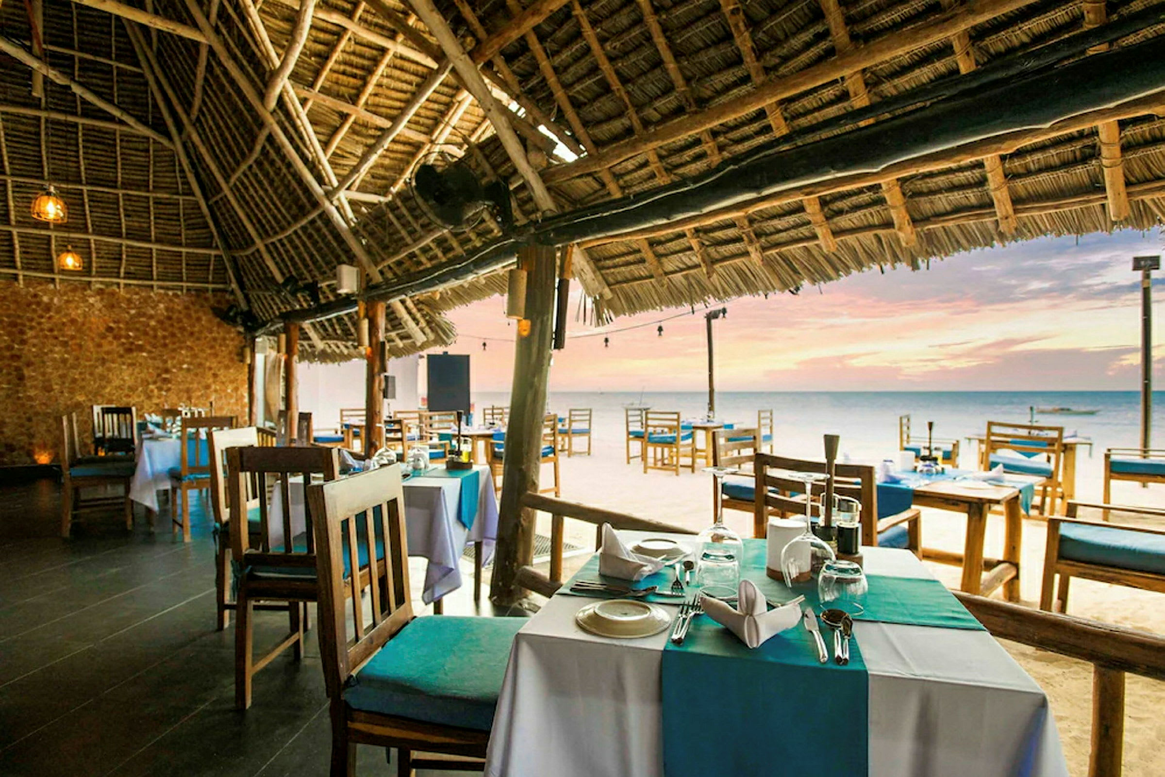 Bilde av en strandnær restaurant med bord og stoler under et stråtak. Havet og solnedgangen er synlig i bakgrunnen. (AI-generert tekst)