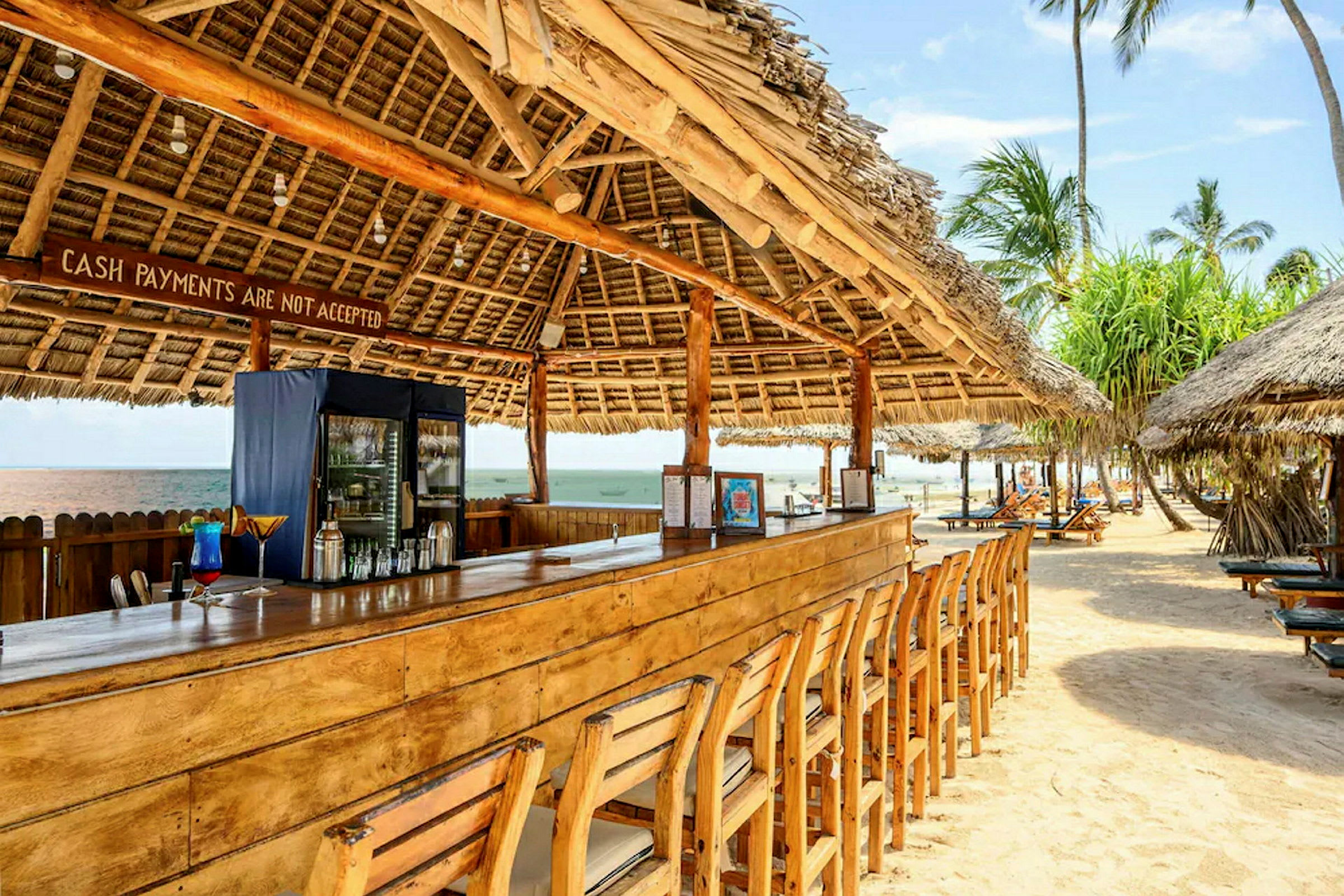 Strandbar med trebord og stoler under tak laget av palmeblader. (AI-generert tekst)