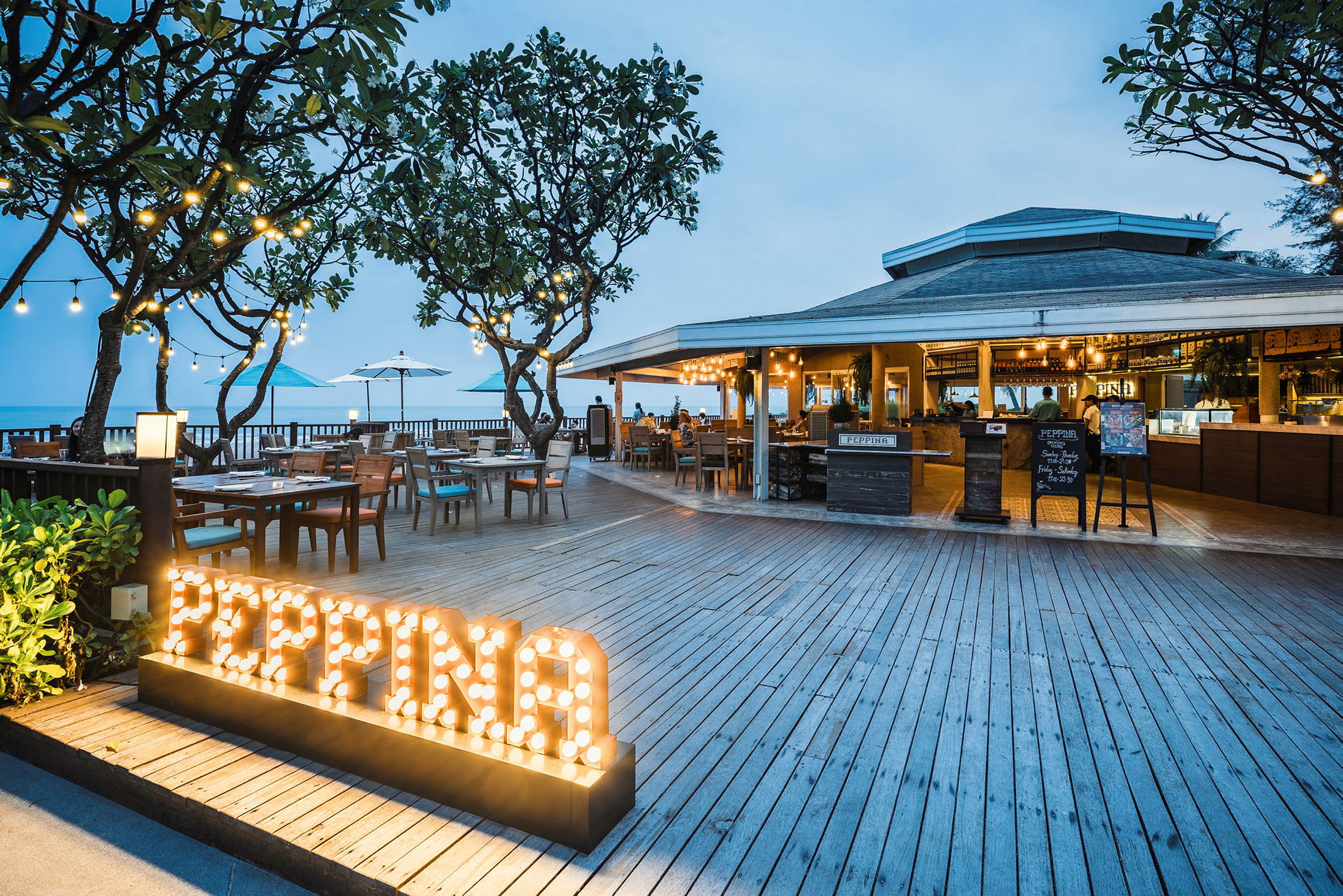 En restaurant med tredekke, bord og sitteplasser utendørs nær sjøen. Skilt med opplyst teksten 'PEPPINA'. (AI-generert tekst)