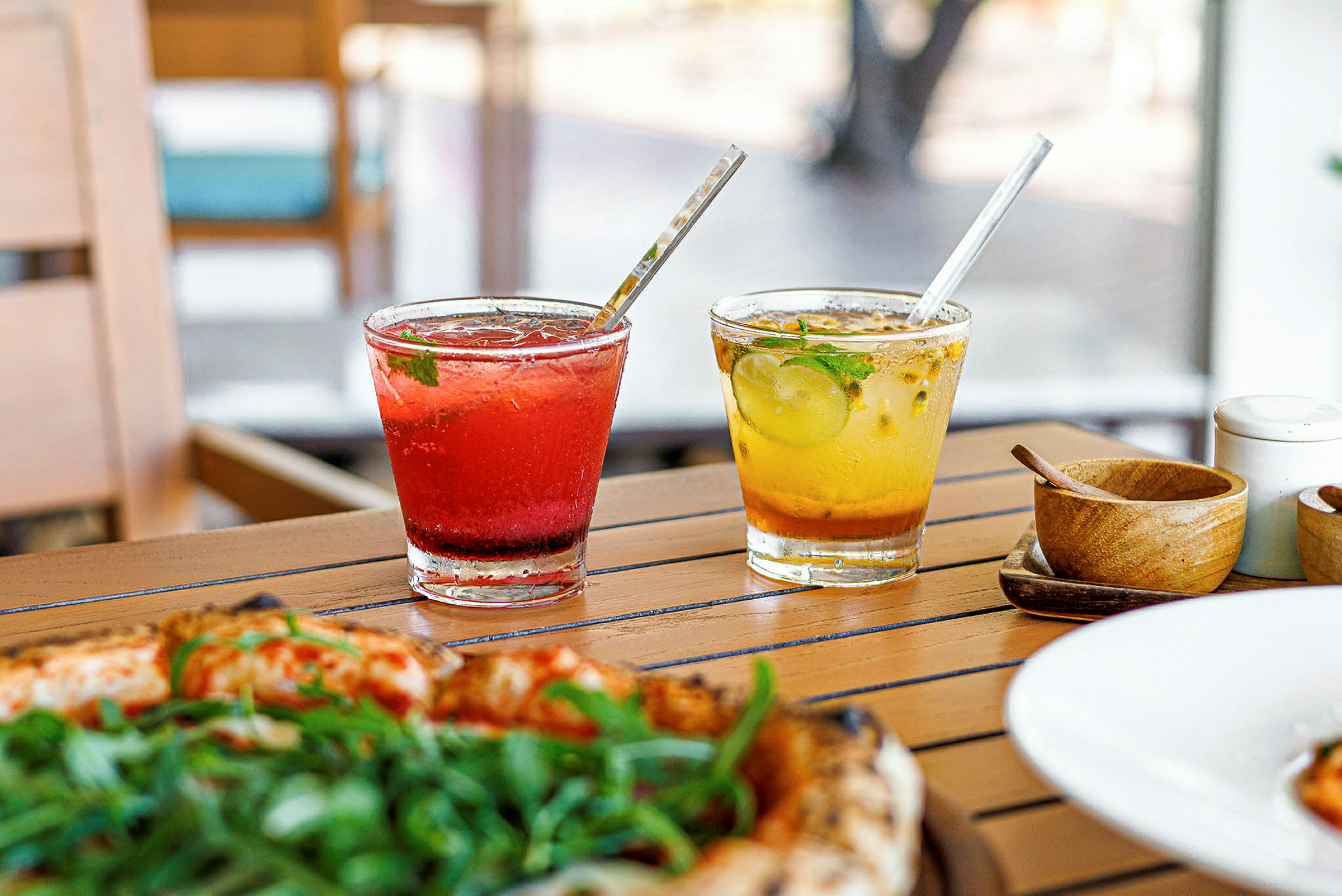 To glass med fargerike drikker står på et trebord. Ett glass inneholder rød drikk, og det andre glasset gul drikk. Foran glassene er en pizza med grønn topping. (AI-generert tekst)