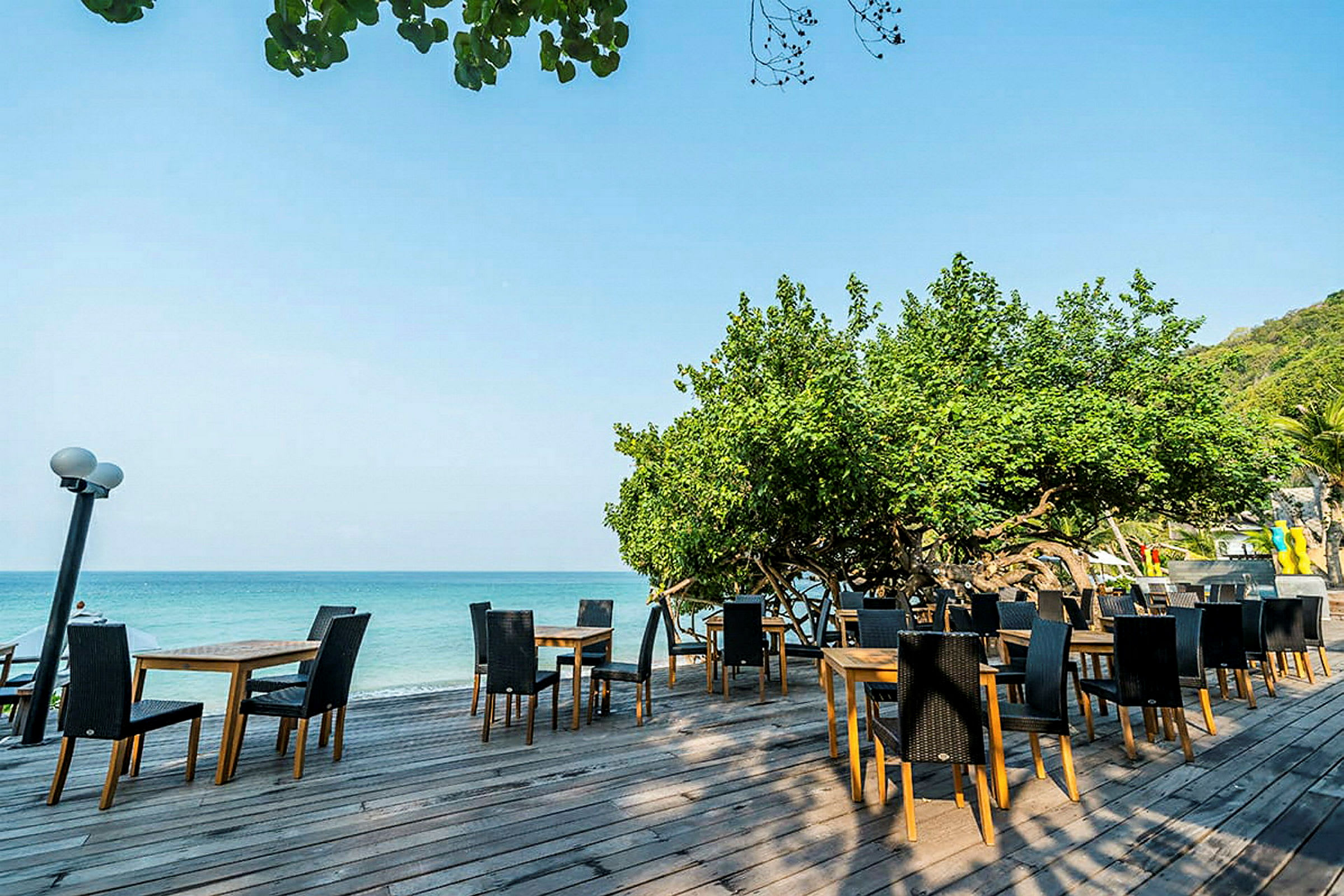 En utendørsrestaurant med bord og stoler plassert på en terrasse av tre ved havet, omgitt av grøntområder. (AI-generert tekst)